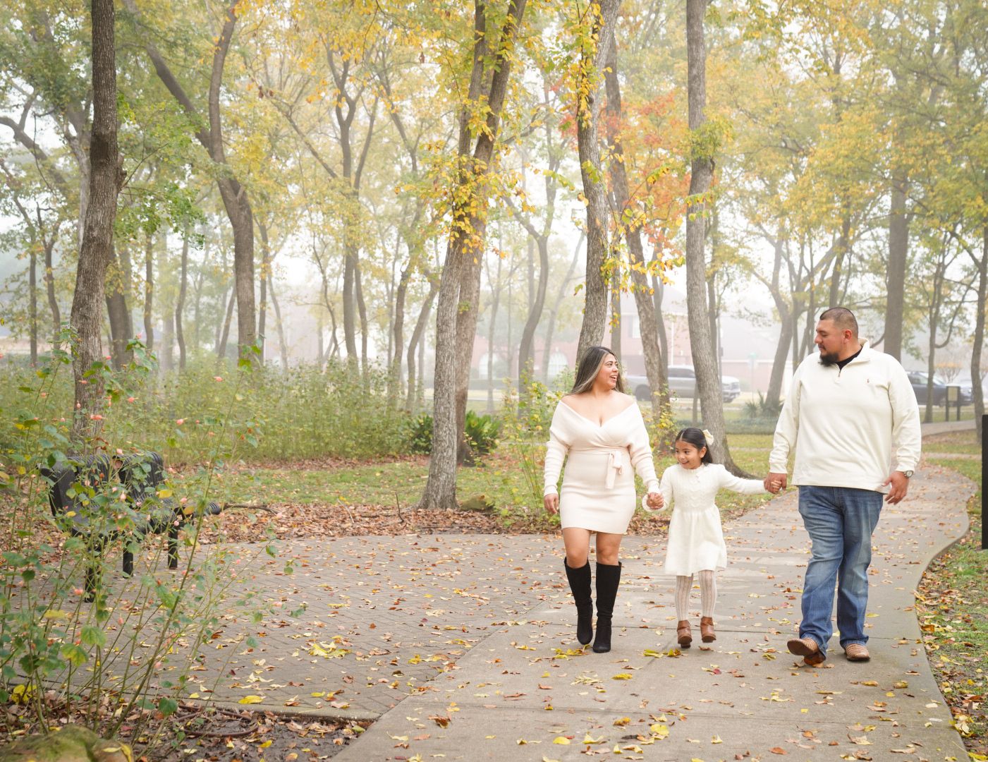 Family of three walking hand-in-hand on a foggy autumn park path surrounded by colorful fall foliage.