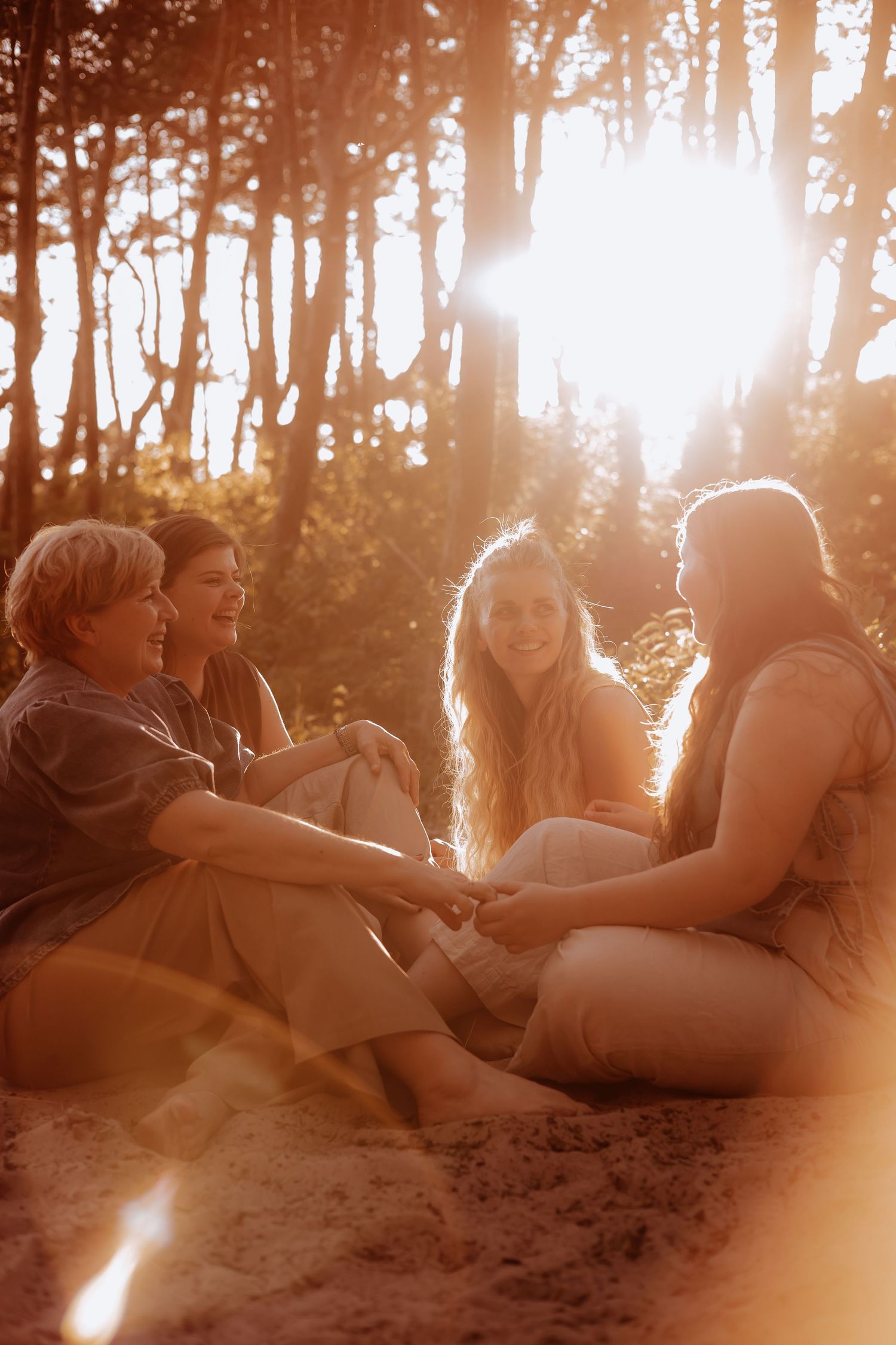 Three women sit together on sandy ground in a glowing forest, laughing and enjoying each other's company.