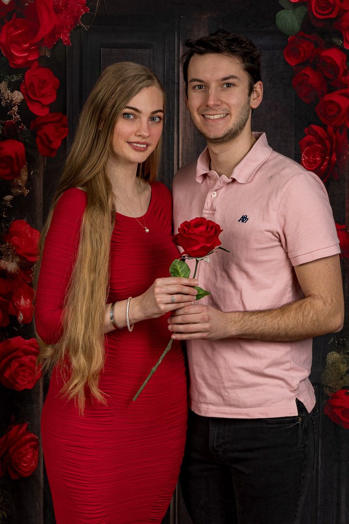 Couple posing with a red rose in front of a floral backdrop, woman in red dress, man in pink polo shirt.