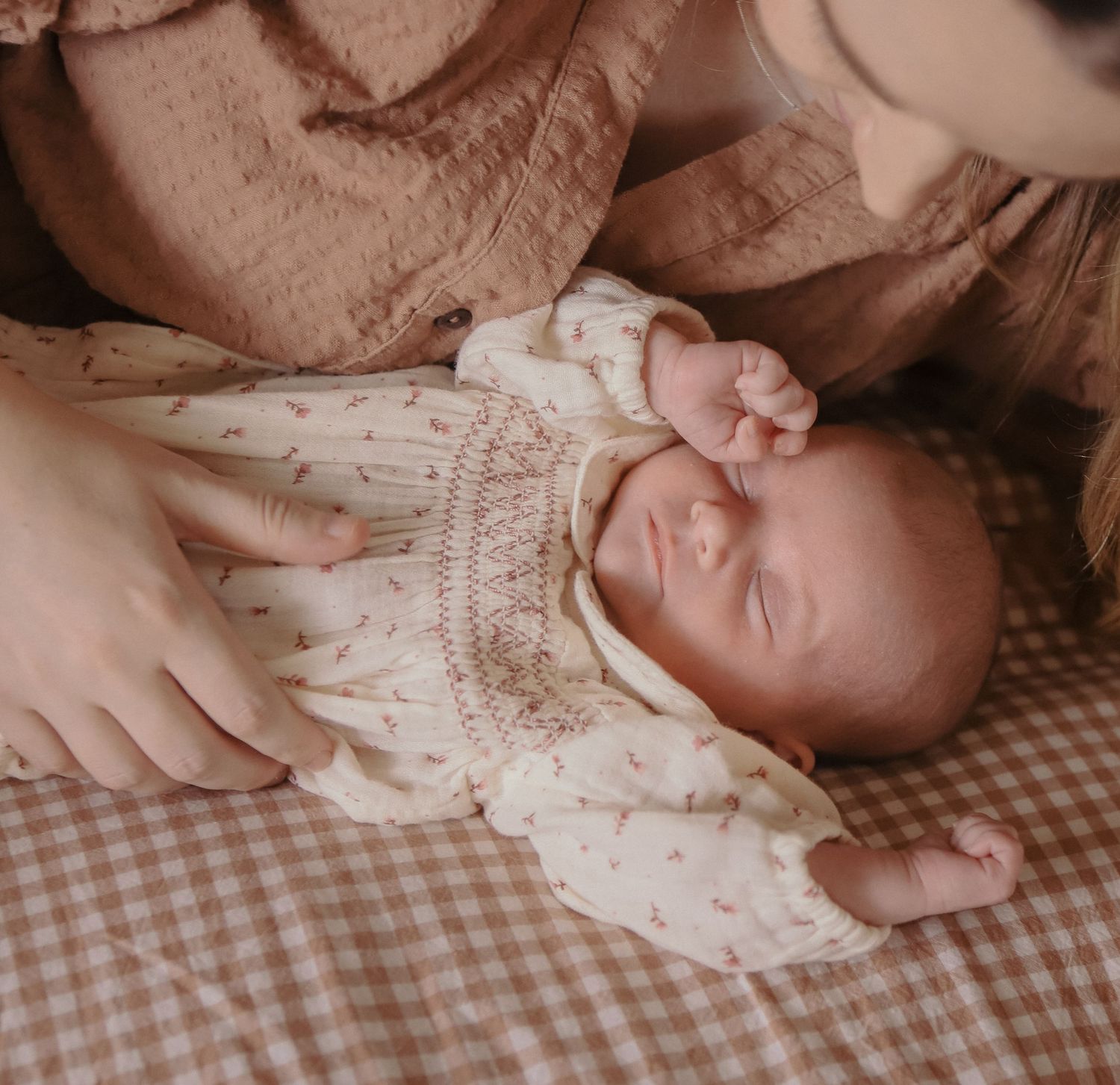 Newborn baby sleeping peacefully in a floral romper on a gingham blanket while a parent gently touches them.