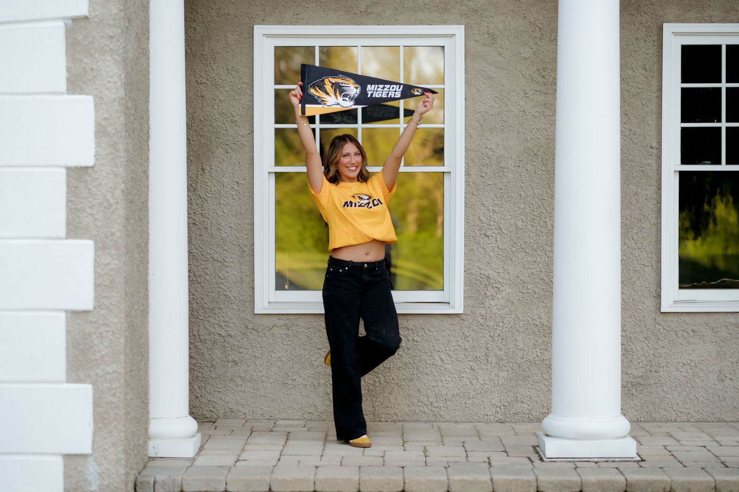Young woman in yellow Mizzou shirt celebrates outdoors, holding a pennant flag while posing by a white-columned building.