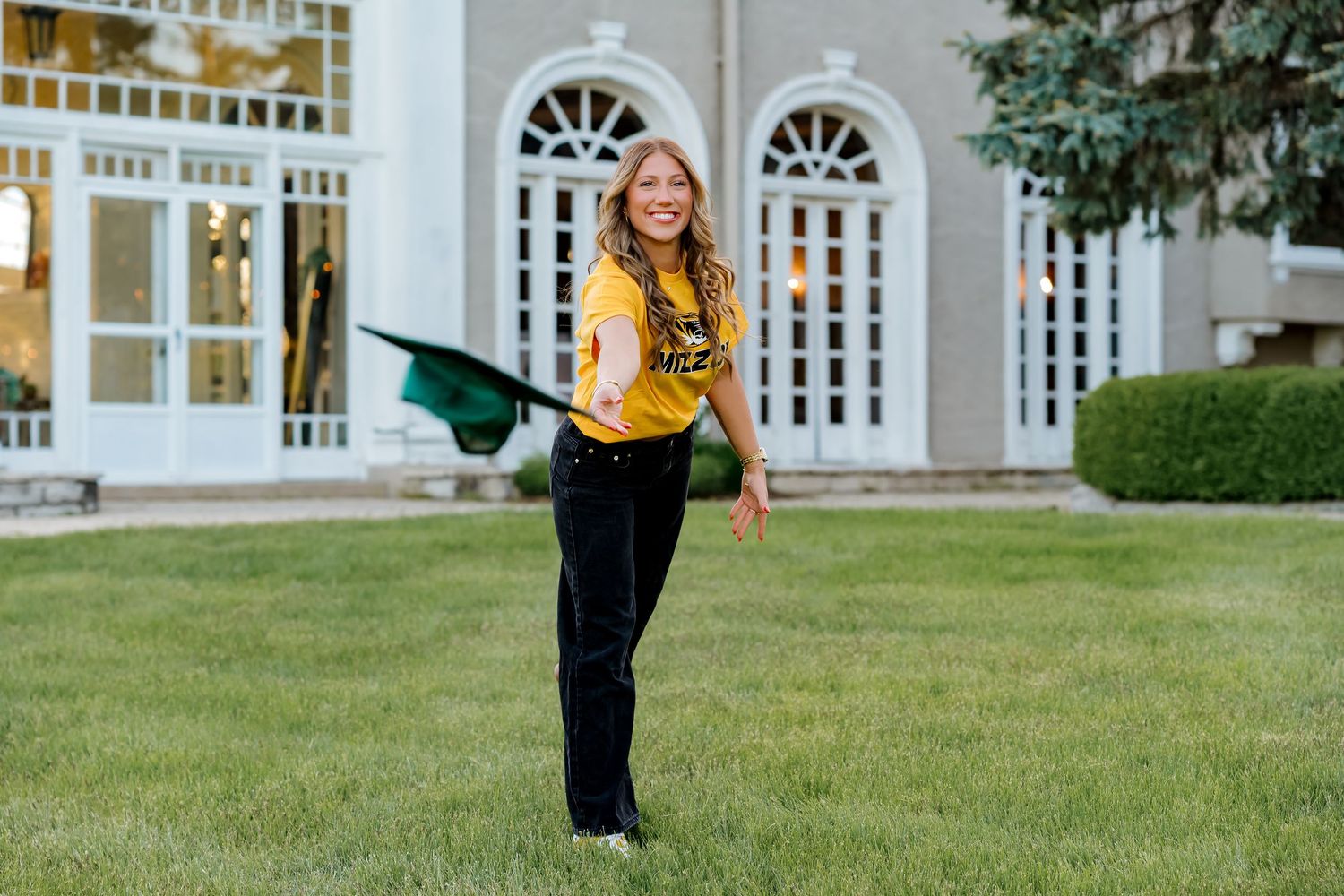 Young woman in gold shirt tossing green graduation cap on lawn in front of elegant white building.