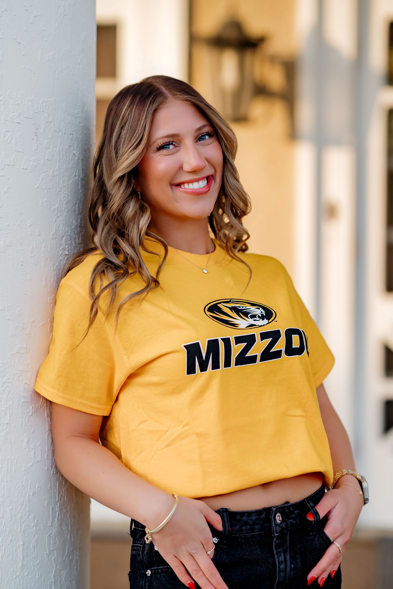 Smiling young woman wearing a gold Mizzou crop top and dark jeans, leaning against a white pillar.