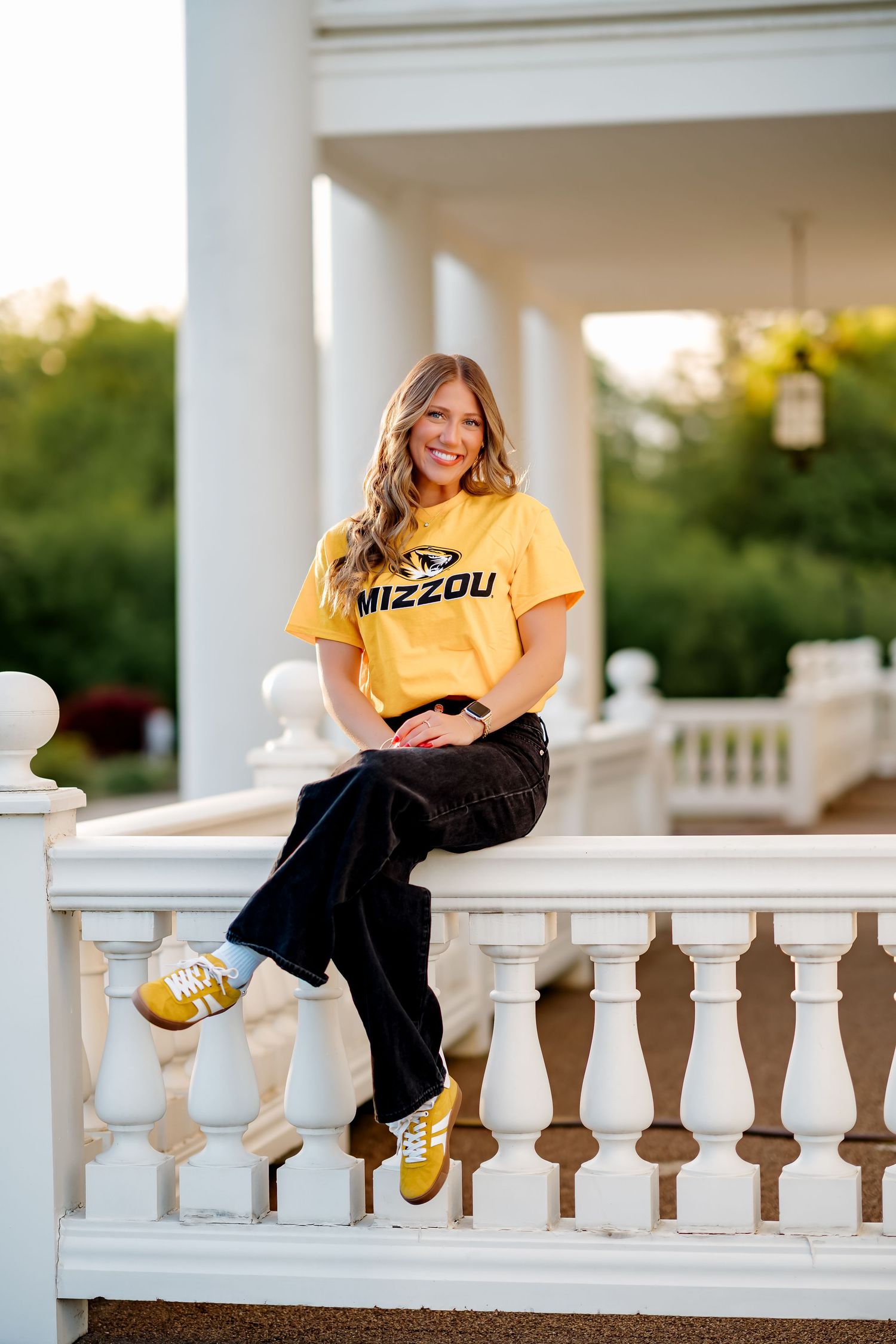 Young woman in a gold Mizzou t-shirt and dark wide-leg jeans sitting on a white balustrade outdoors.