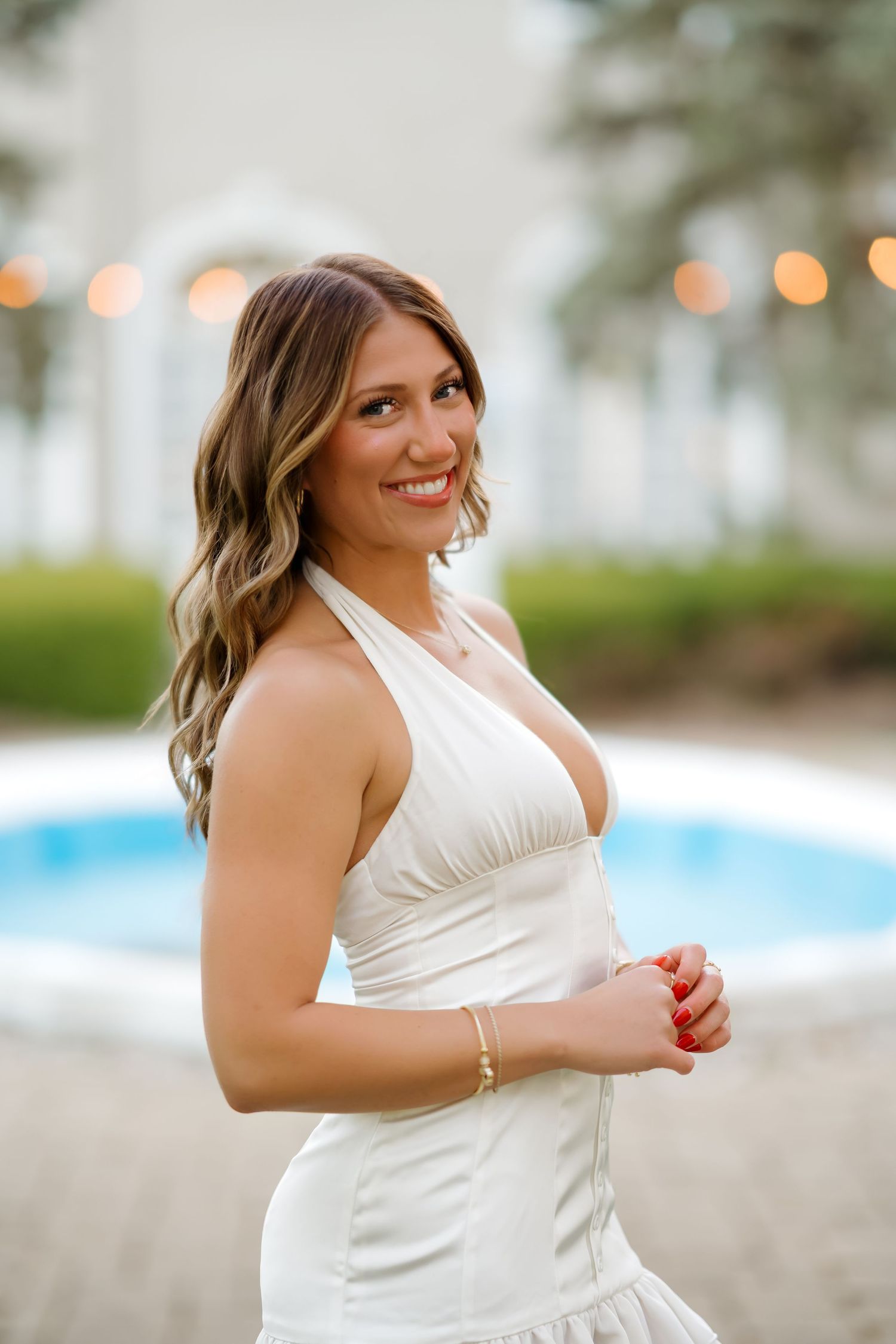 Woman in white halter dress smiling by pool with string lights.