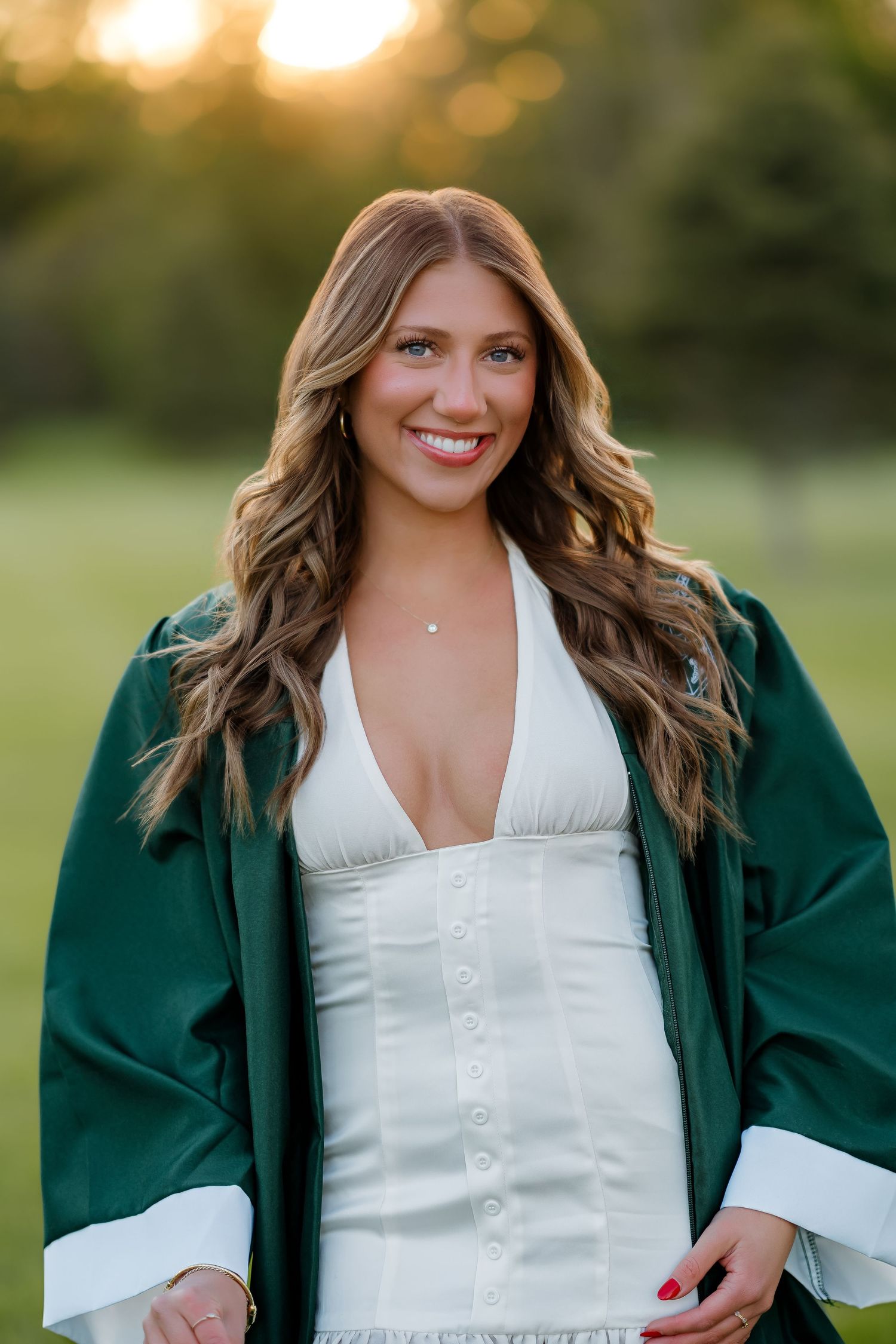 Smiling graduate poses outdoors in white dress and green graduation robe at golden hour sunset.