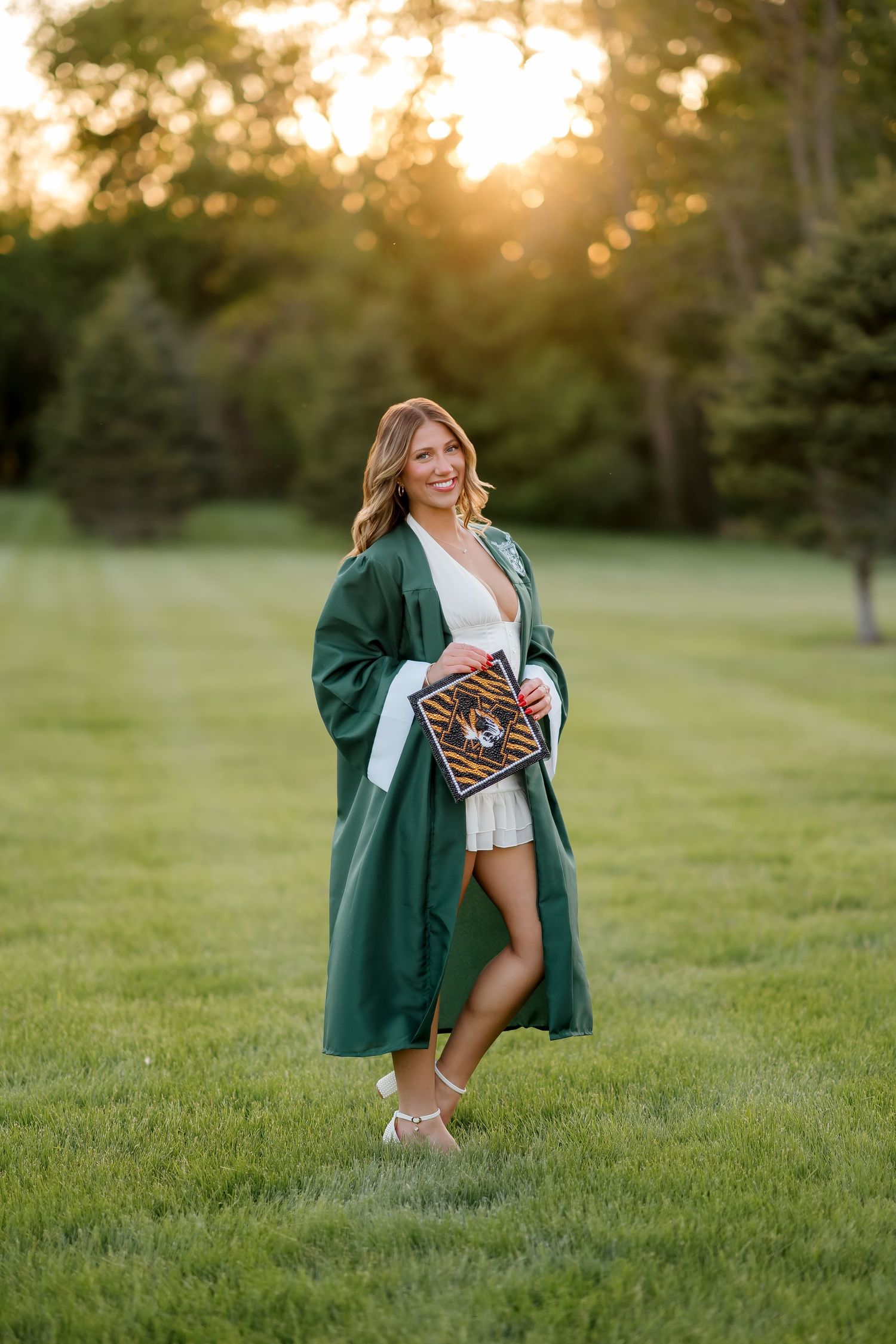 Young woman in green graduation gown holding cap, smiling outdoors at golden hour sunset on green lawn.