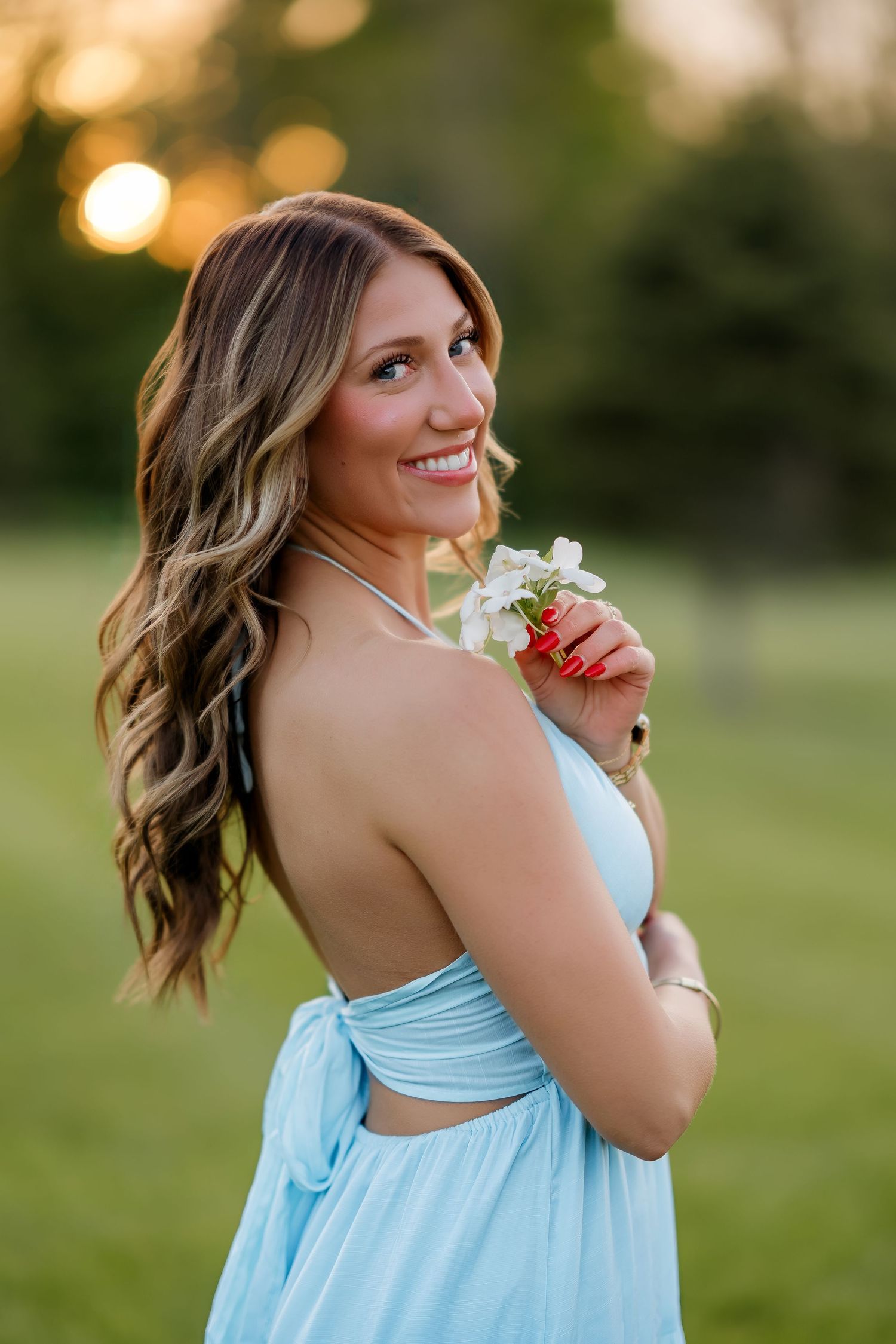 Smiling woman in backless light blue halter maxi dress holding white flowers at golden hour outdoors.