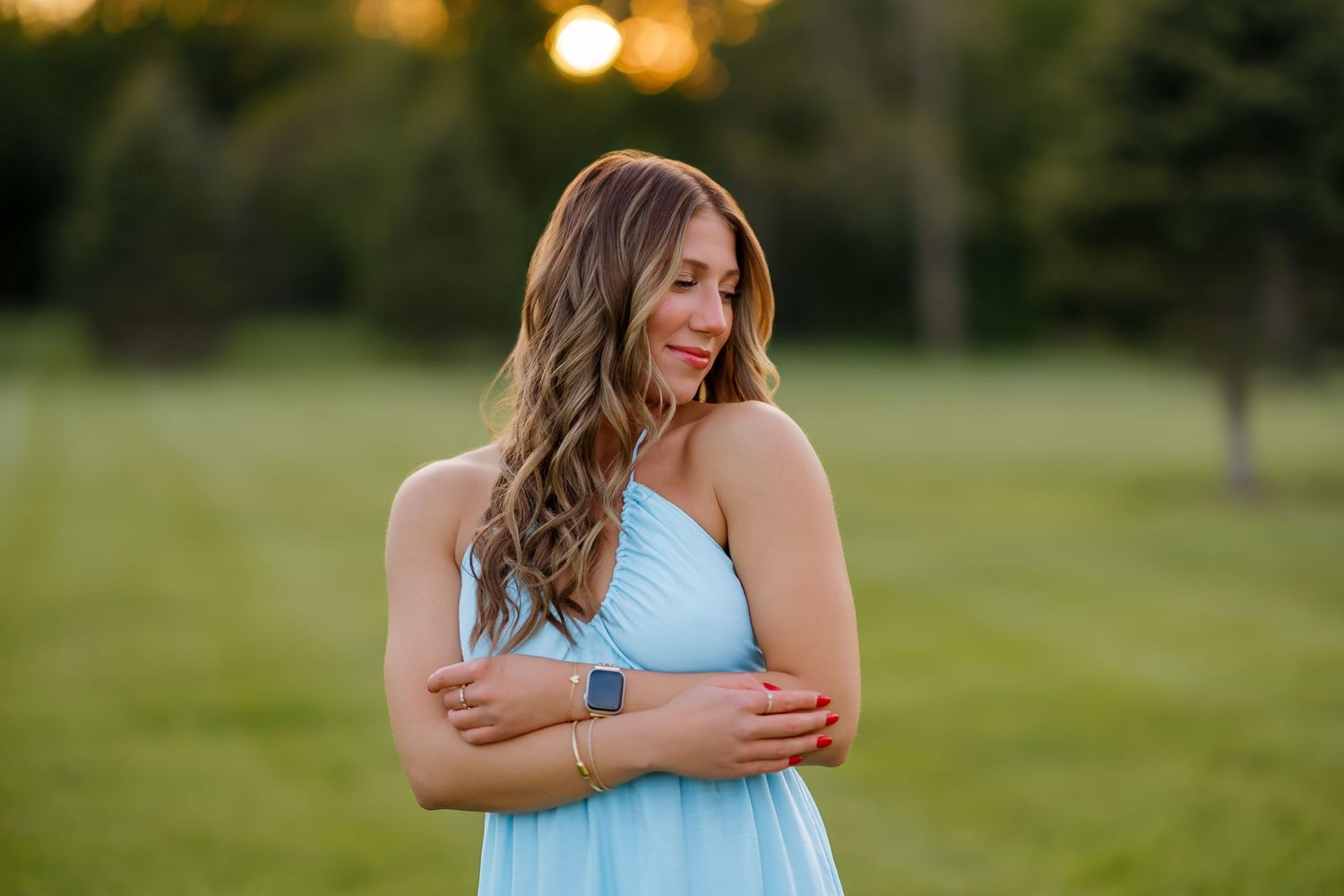 Young woman in light blue halter dress with arms crossed smiling softly at golden hour in green field.