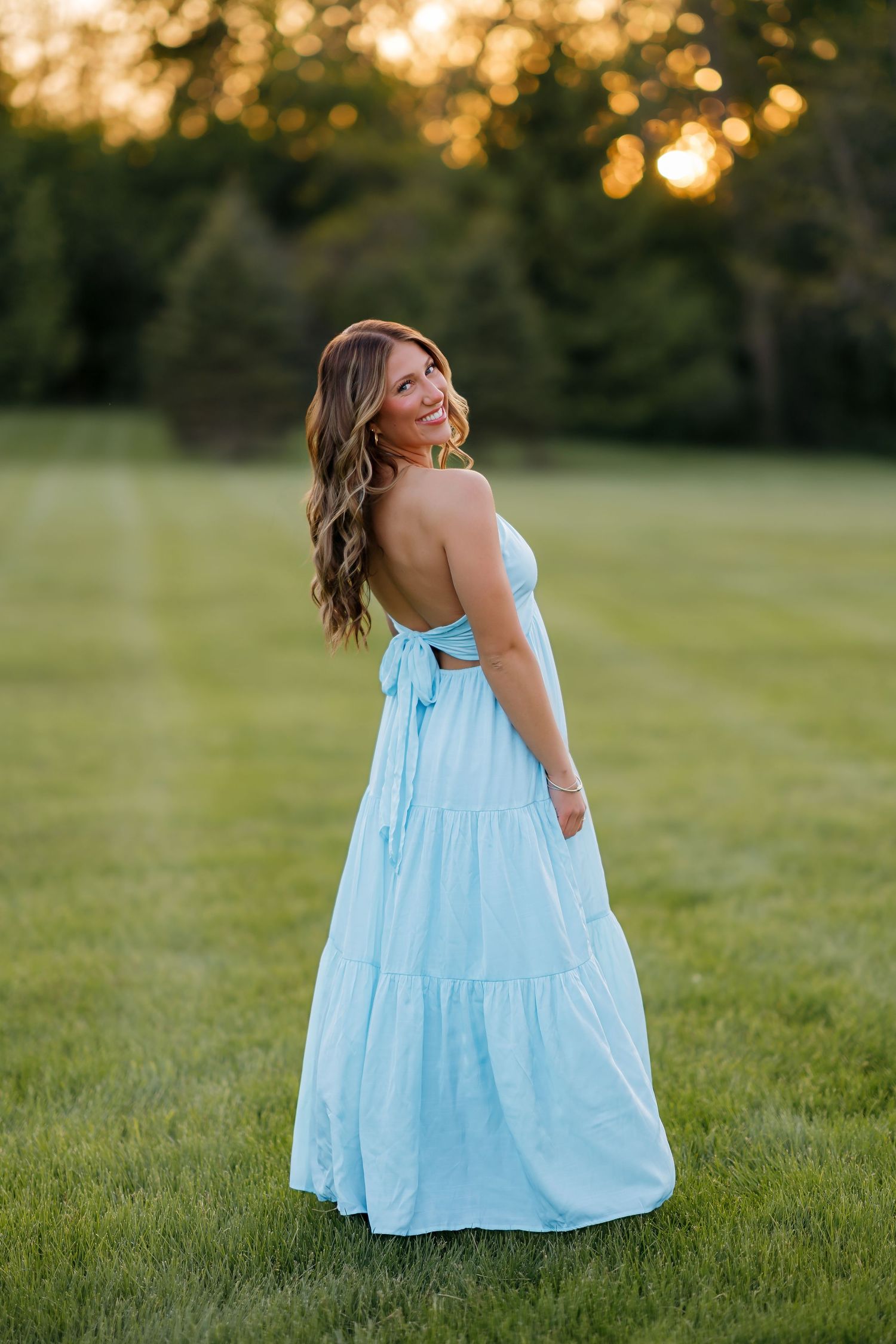 Woman in backless light blue tiered maxi dress smiling over shoulder at sunset in lush green field.
