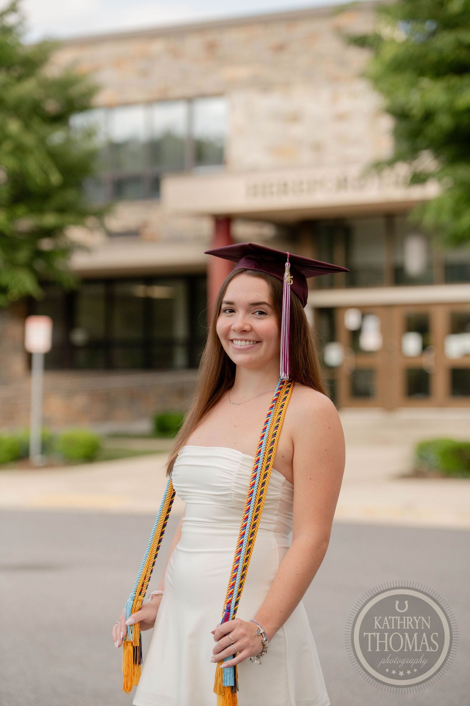 Graduate in white dress and cap poses outdoors with honor cords during university commencement ceremony.