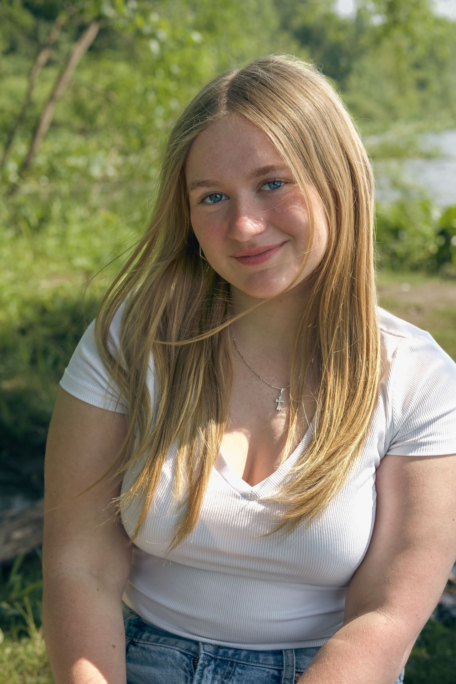 A person in a white t-shirt sits outdoors on a sunny day against a green natural background.