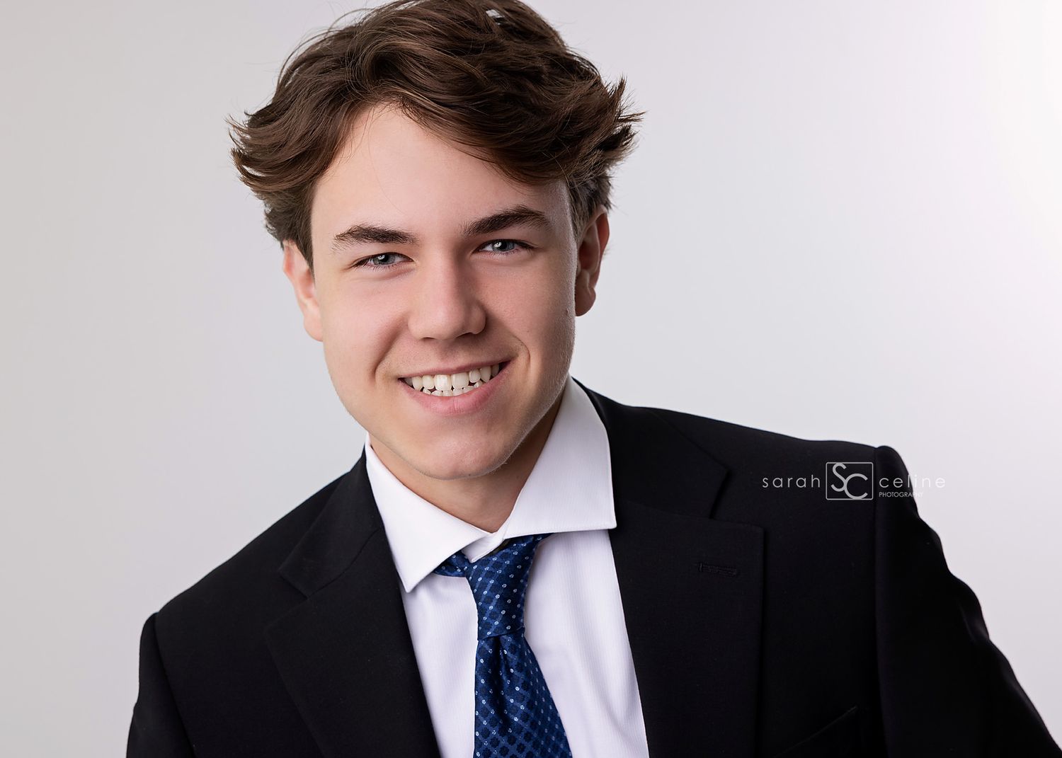 Professional headshot of a person in a dark suit jacket, white dress shirt and blue tie with styled brown hair.