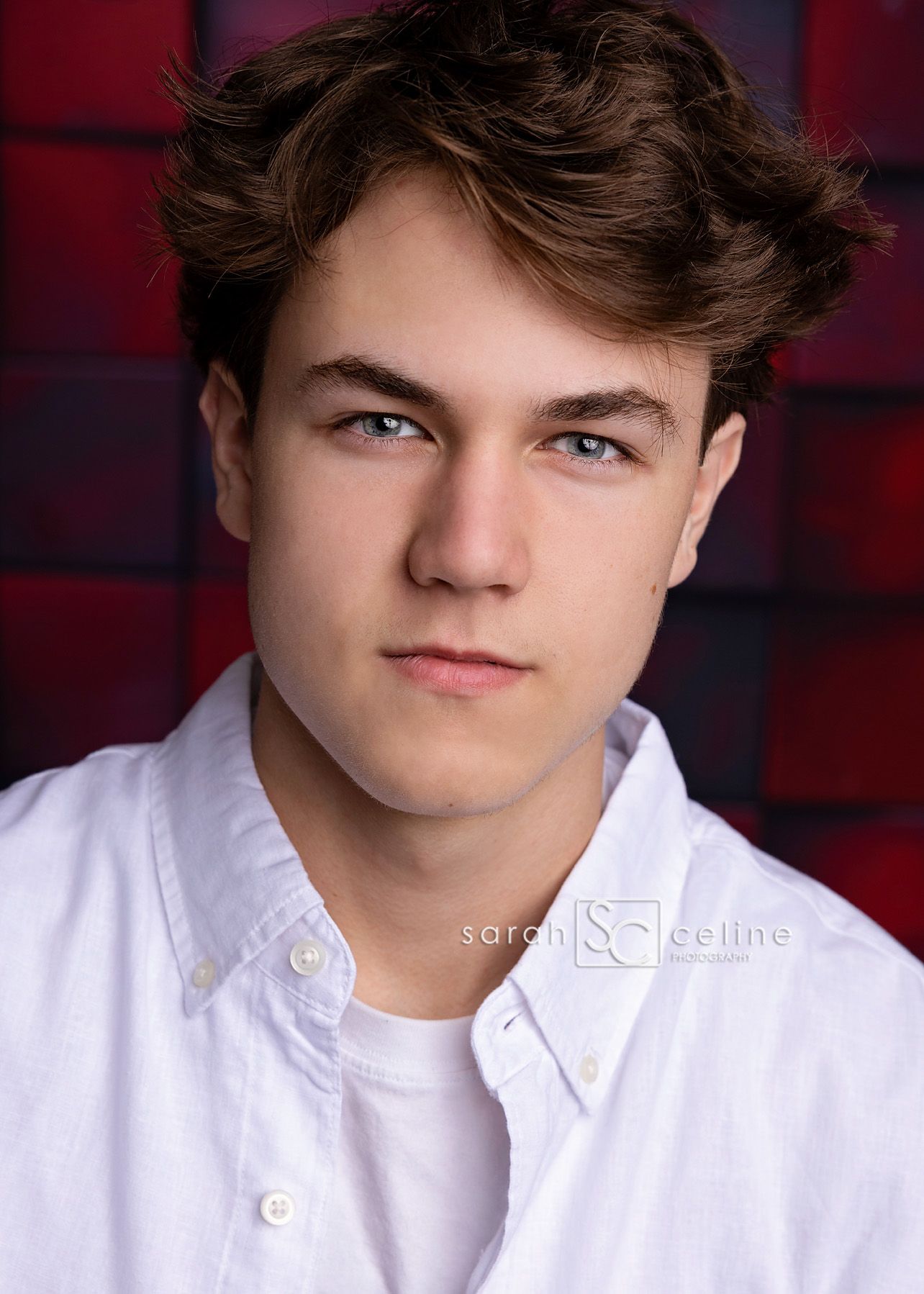 Young person wearing a white button-down shirt poses against a dark red backdrop in a professional headshot portrait.