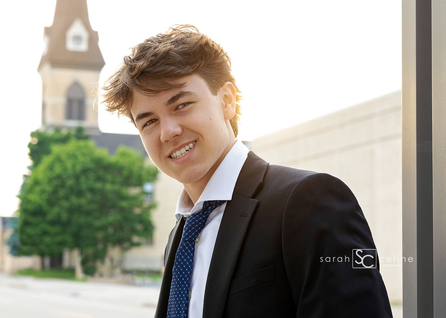 A young graduate in a black suit and blue tie stands outdoors with a clock tower visible in the background at sunset.