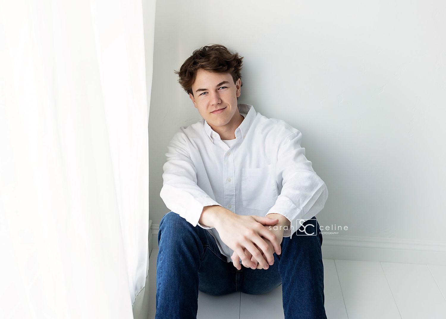 Camera portrait of someone wearing a white collared shirt and blue jeans sitting against a light wall.