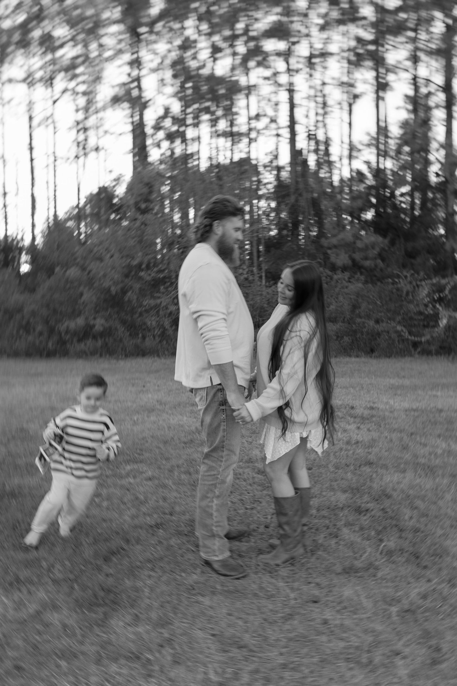 A couple holds hands in a pine forest field while a young child runs joyfully toward them in black and white.