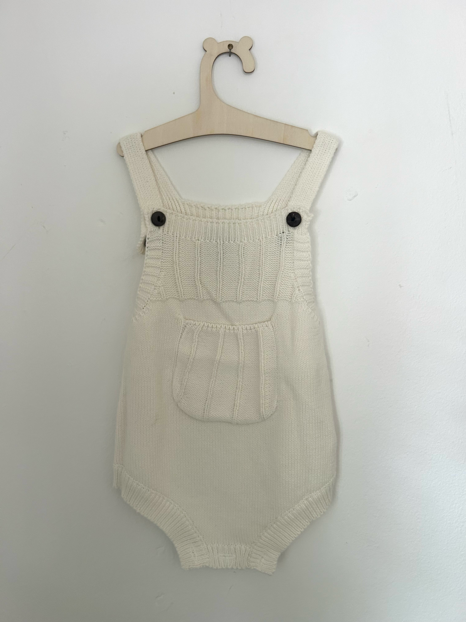 Vintage-style beige knitted baby romper with button details and side pockets hanging on display.