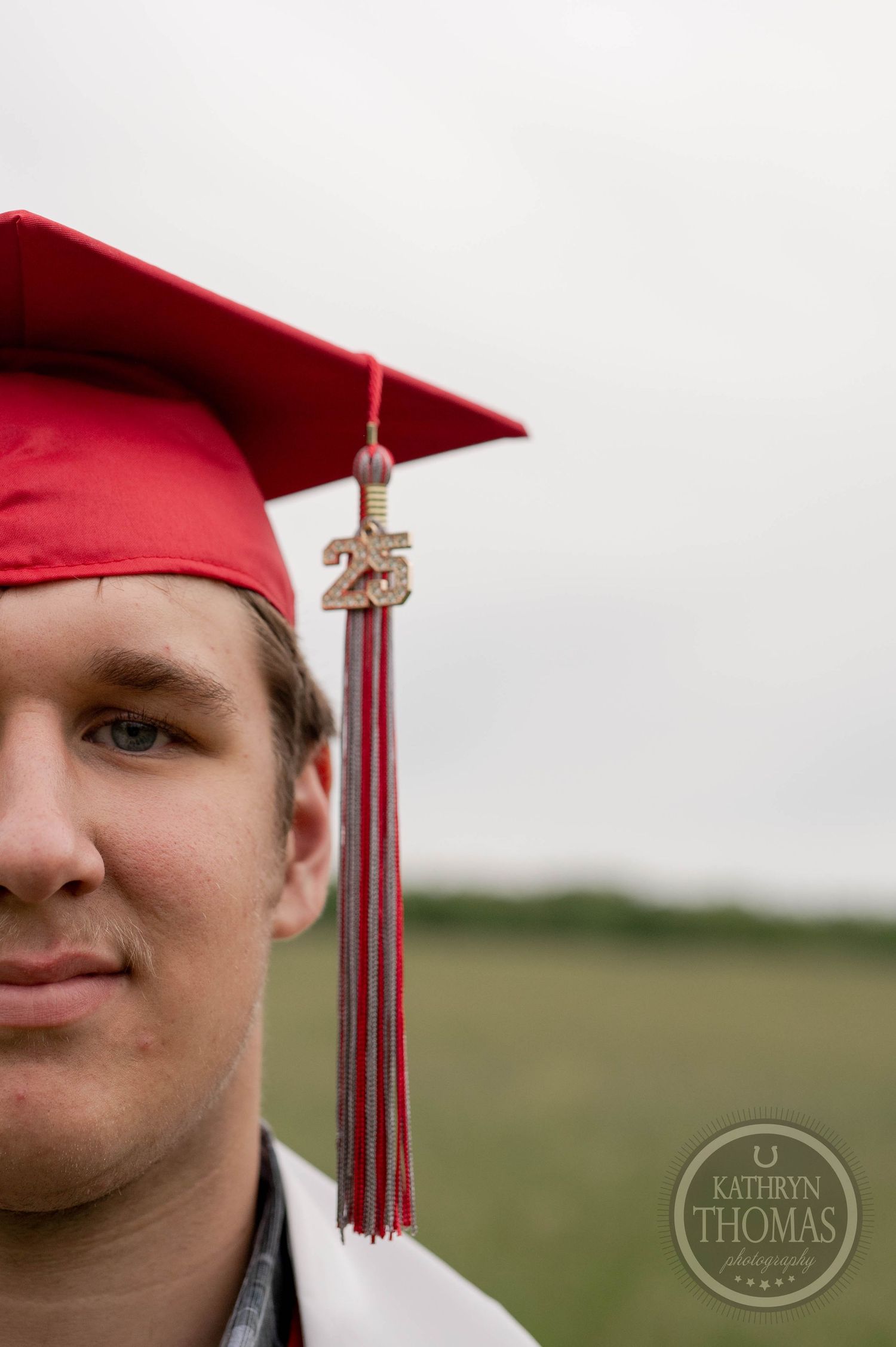 Graduate in red cap and gown with class tassel standing outdoors against a blurred natural background.