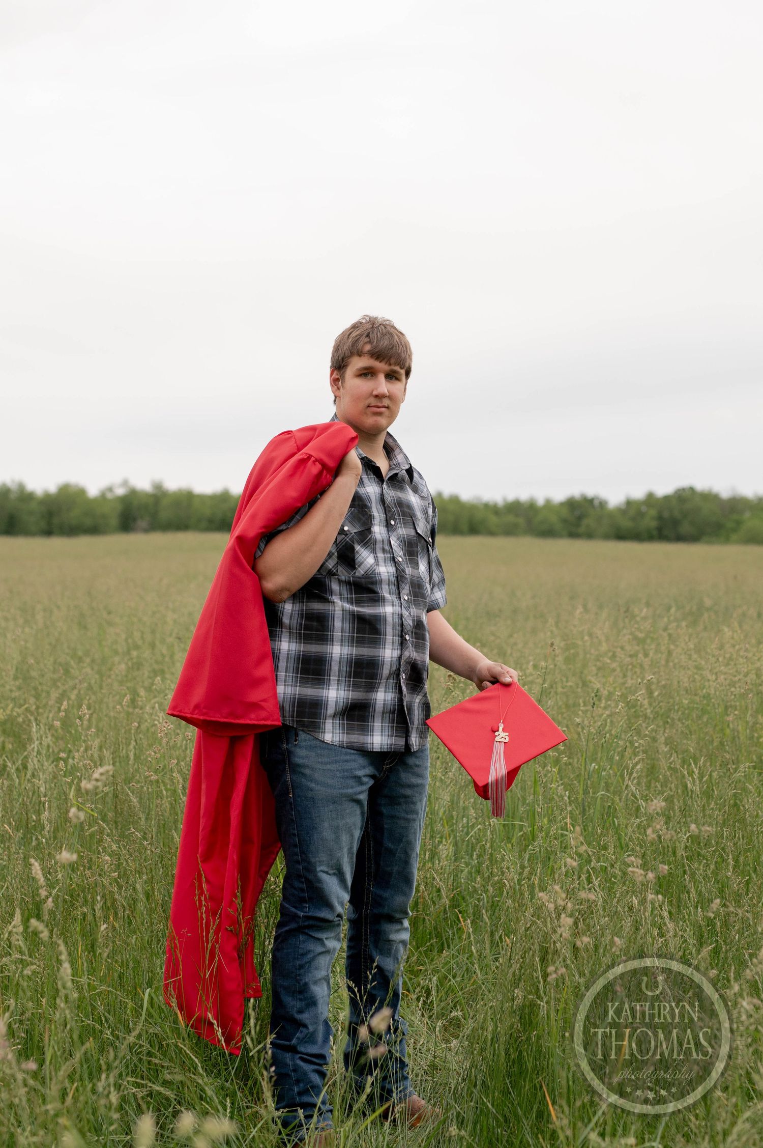 Graduate in plaid shirt and red graduation gown stands in a grassy field holding a red diploma cover.