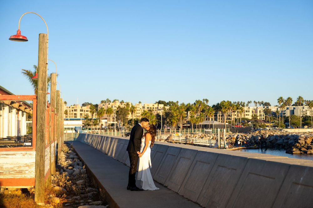 A couple embraces at sunset on a scenic marina boardwalk with palm trees and boats in the background.