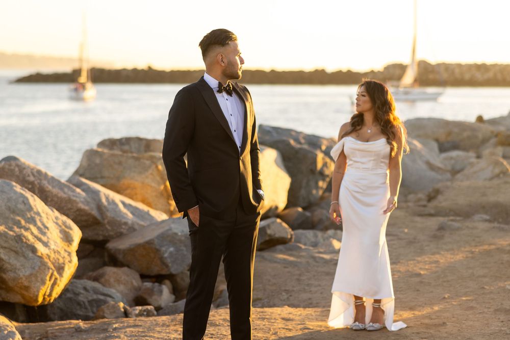 Two people in formal wedding attire stand together on rocky coastal shore at sunset with sailboats in background.