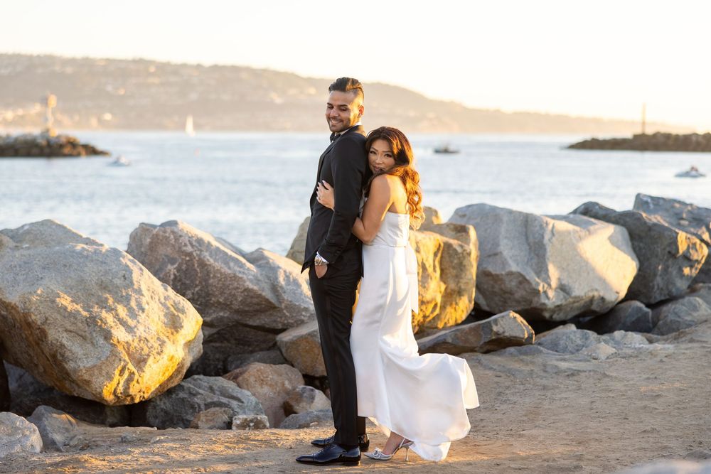 A couple embraces on a rocky beach at sunset with flowing white dress and ocean views in the background.