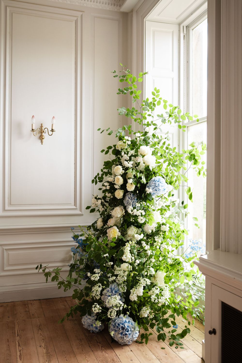 Corner floral arrangement with white roses, hydrangeas and cascading greenery by a window.