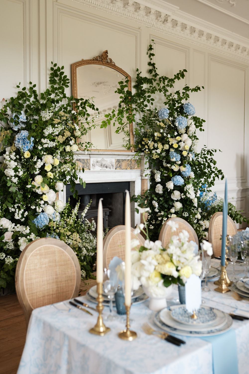Elegant wedding reception table with lush floral arrangements and ornate gold mirror above fireplace.