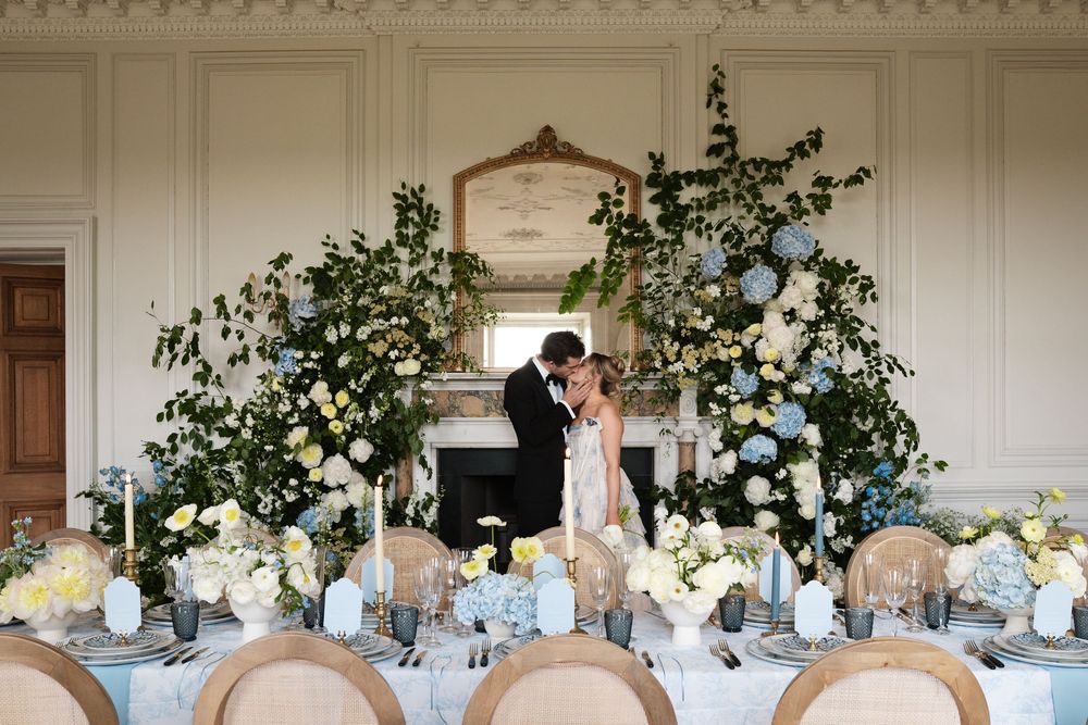 Reception table with floral arch and fireplace backdrop featuring elegant place settings and white flowers.