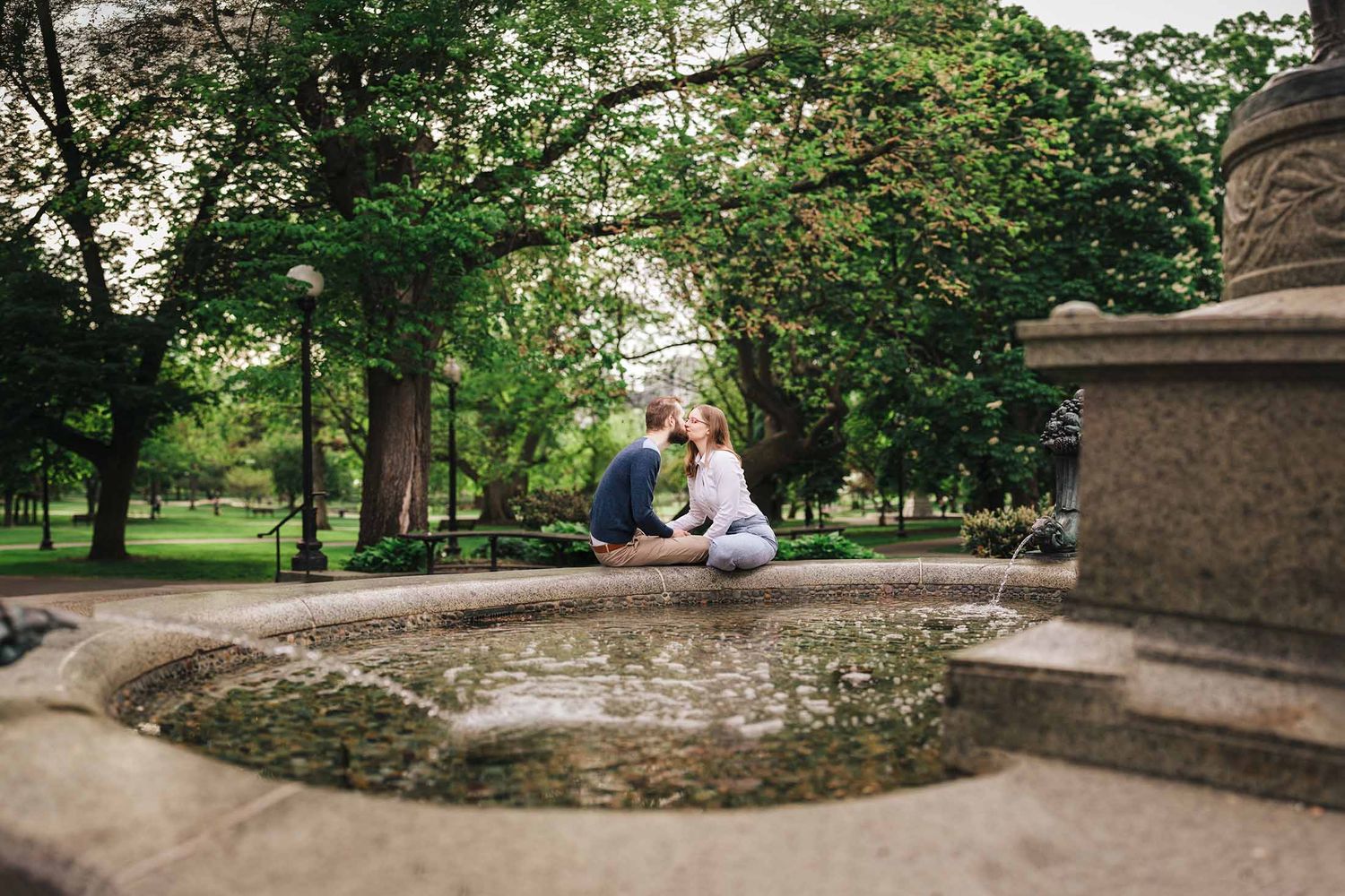 Wide angle engagement portrait of couple kissing beside the George Robert White Memorial Fountain with lush greenery in Boston Public Garden