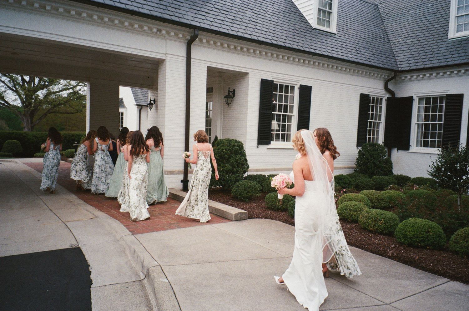 Bridesmaids in floral dresses walk in line outside an elegant white colonial-style house during a wedding ceremony.