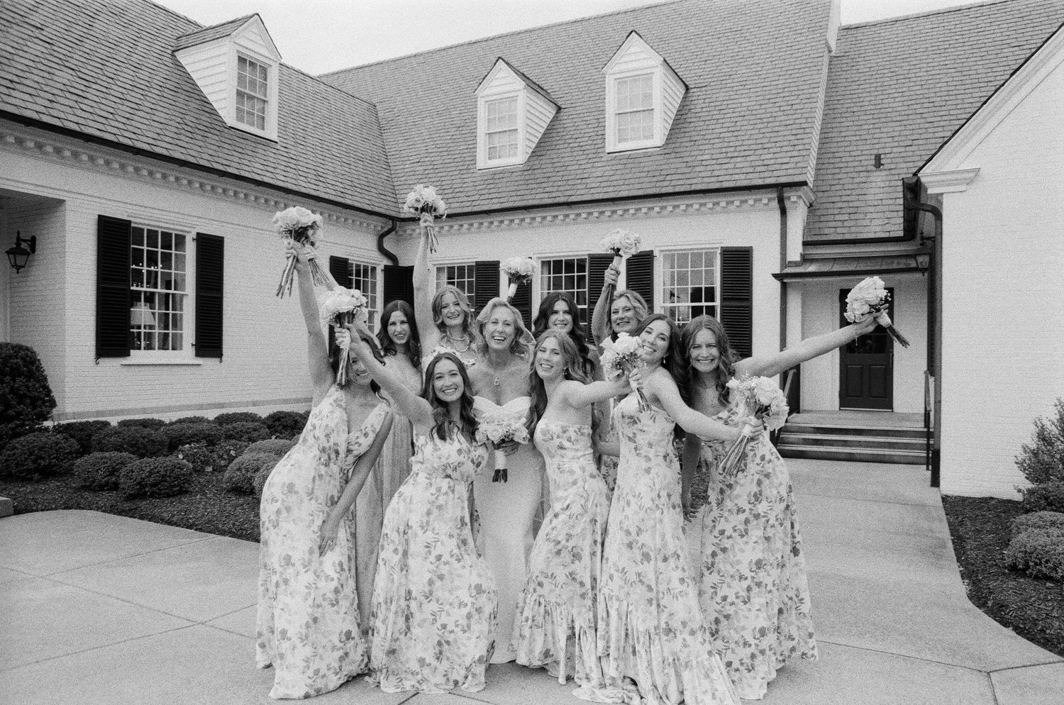 A wedding party in matching floral dresses stands in front of a white Cape Cod-style house.