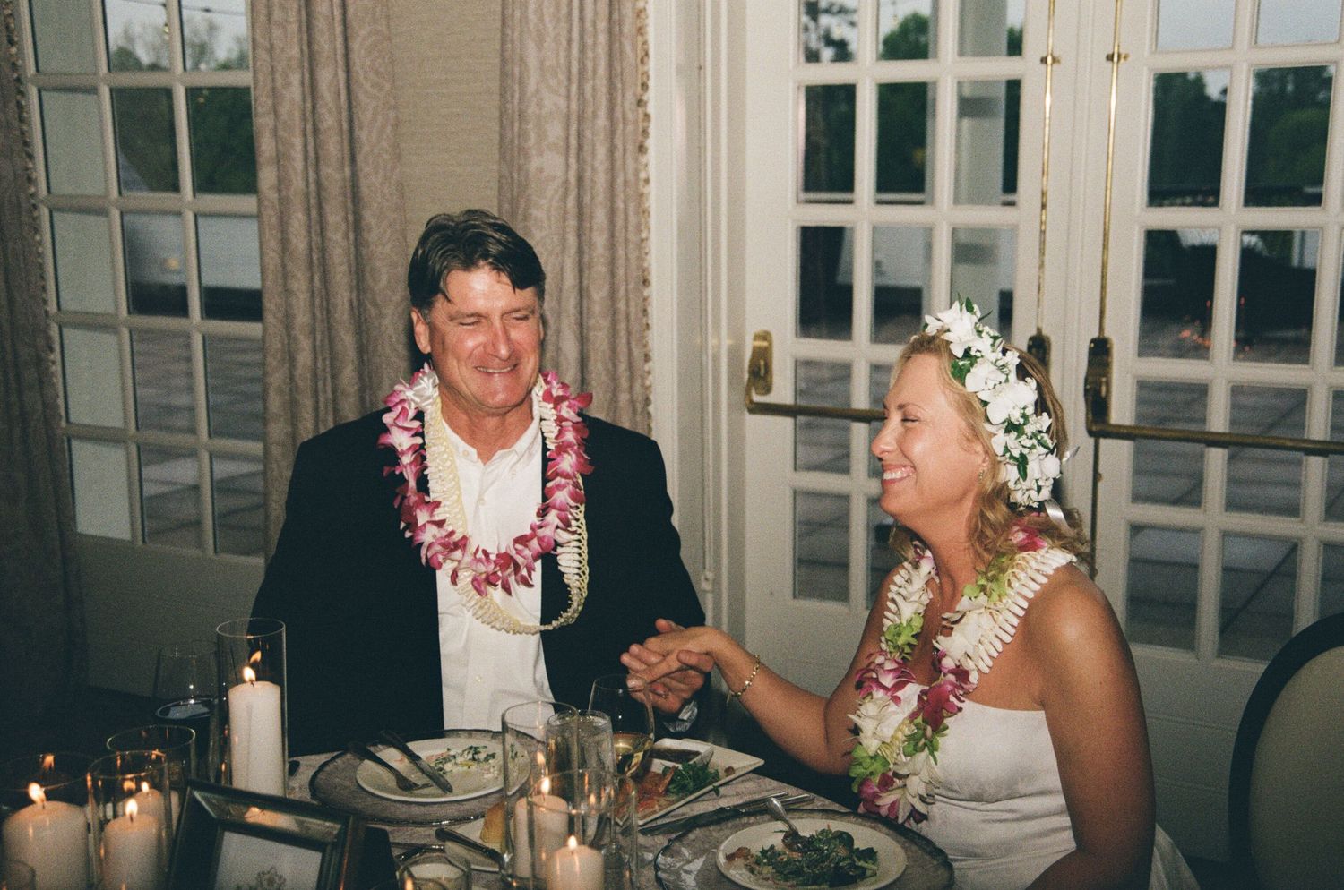 A couple wearing Hawaiian leis dining at an elegant table with candlelight.