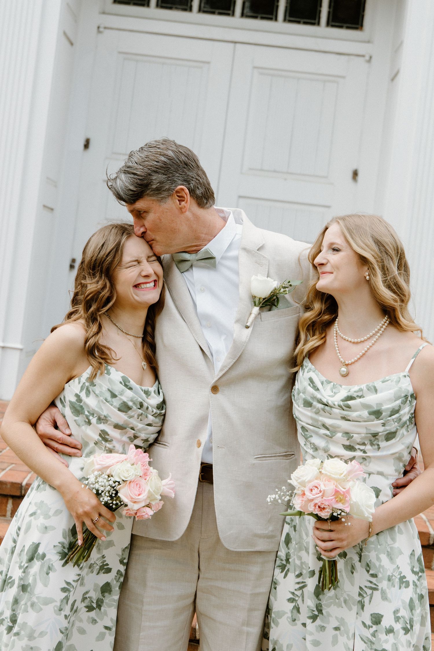 Wedding party members in pale green floral dresses and light beige suit share joyful moment together outside white door entrance.