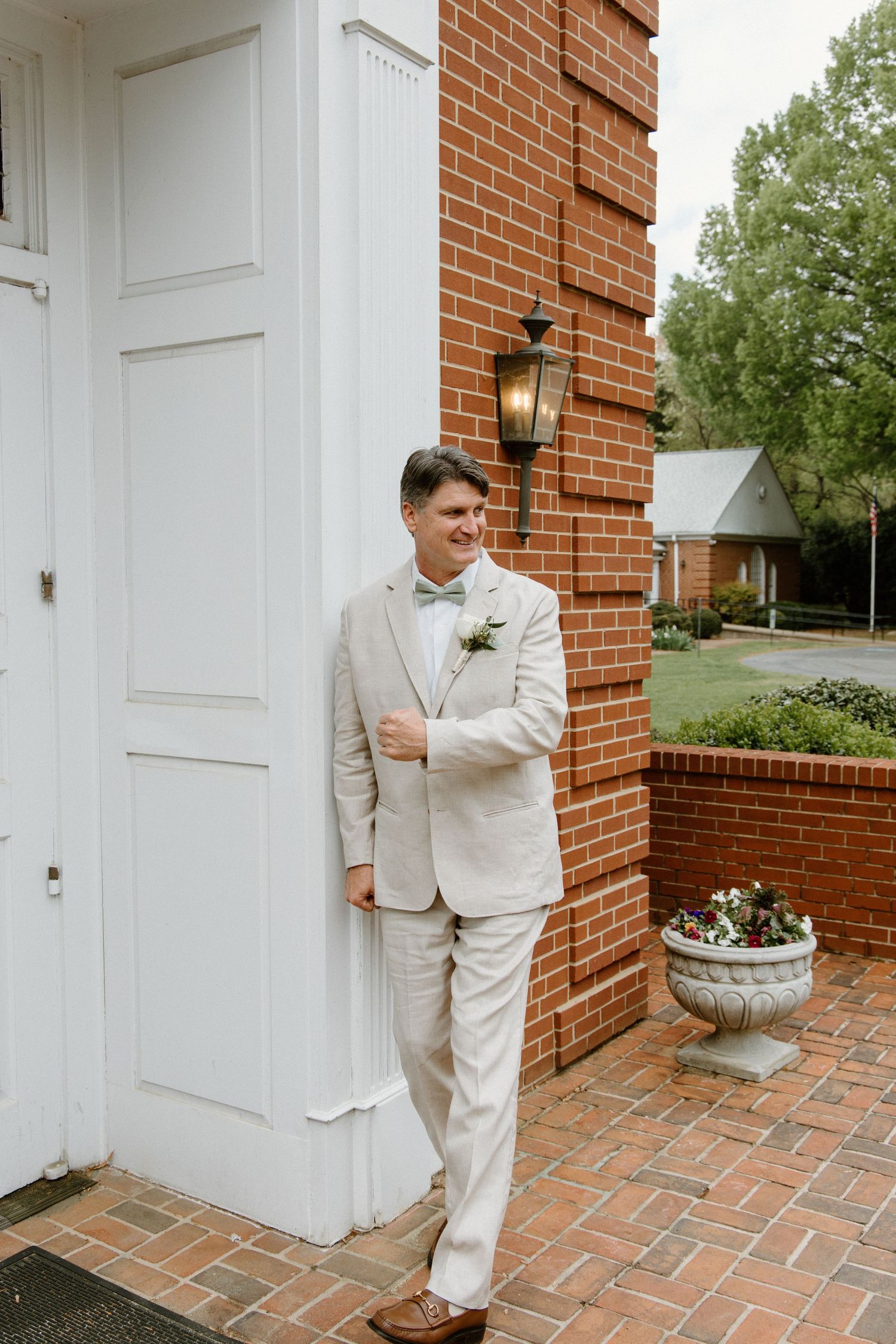 A person in a light beige suit and bow tie poses against a brick wall next to a white door on a brick patio.