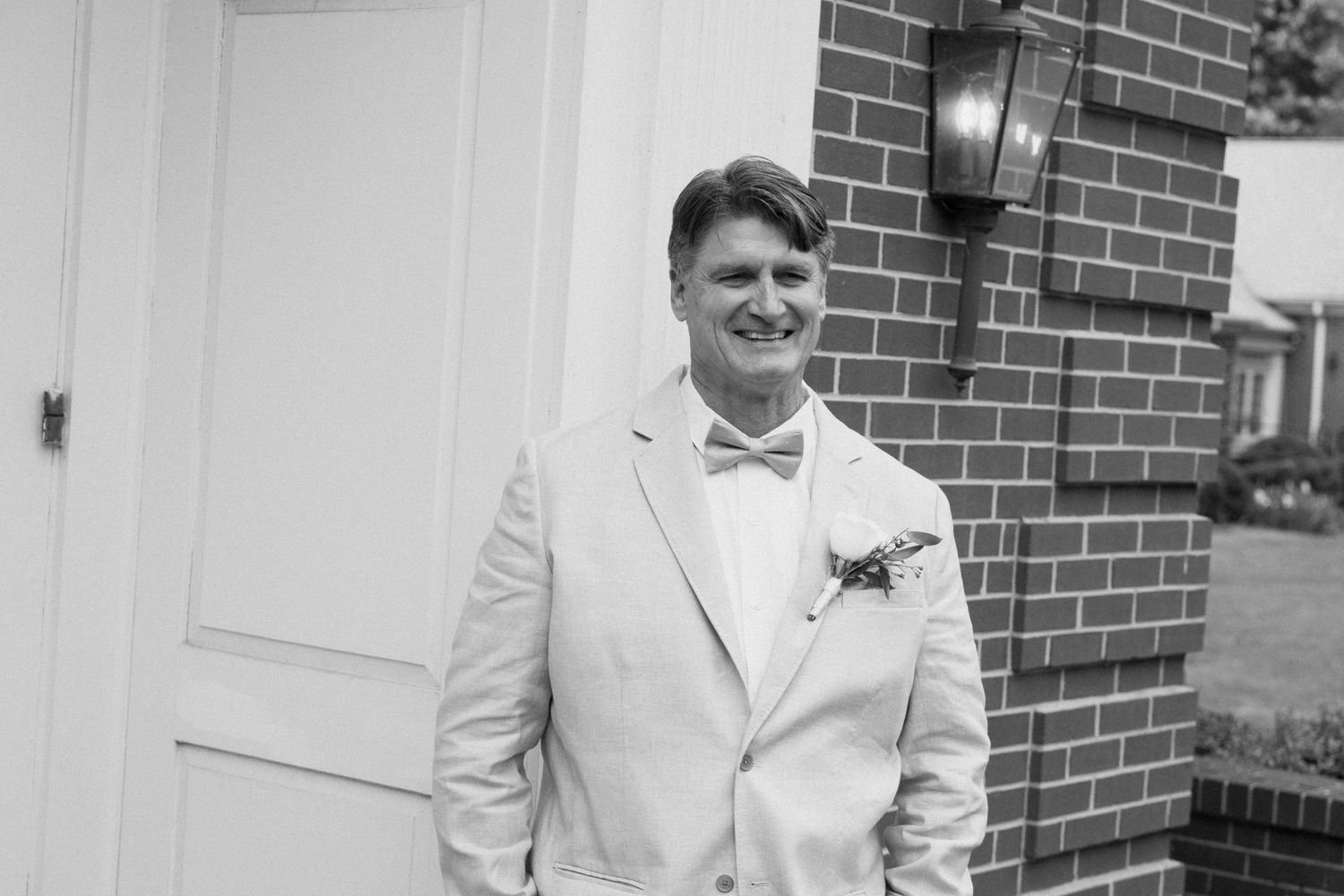 A person in a light colored suit with a bow tie stands smiling against a brick wall exterior.