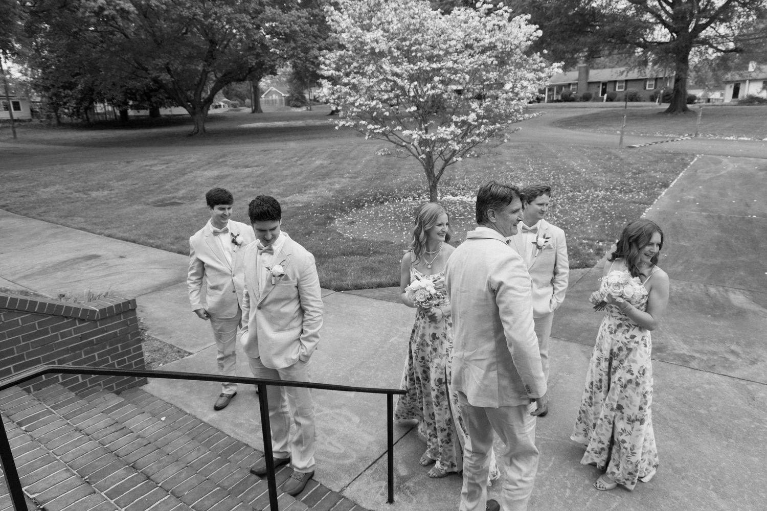 Group in formal attire ascends outdoor stairs by flowering tree at springtime gathering in black and white photo.