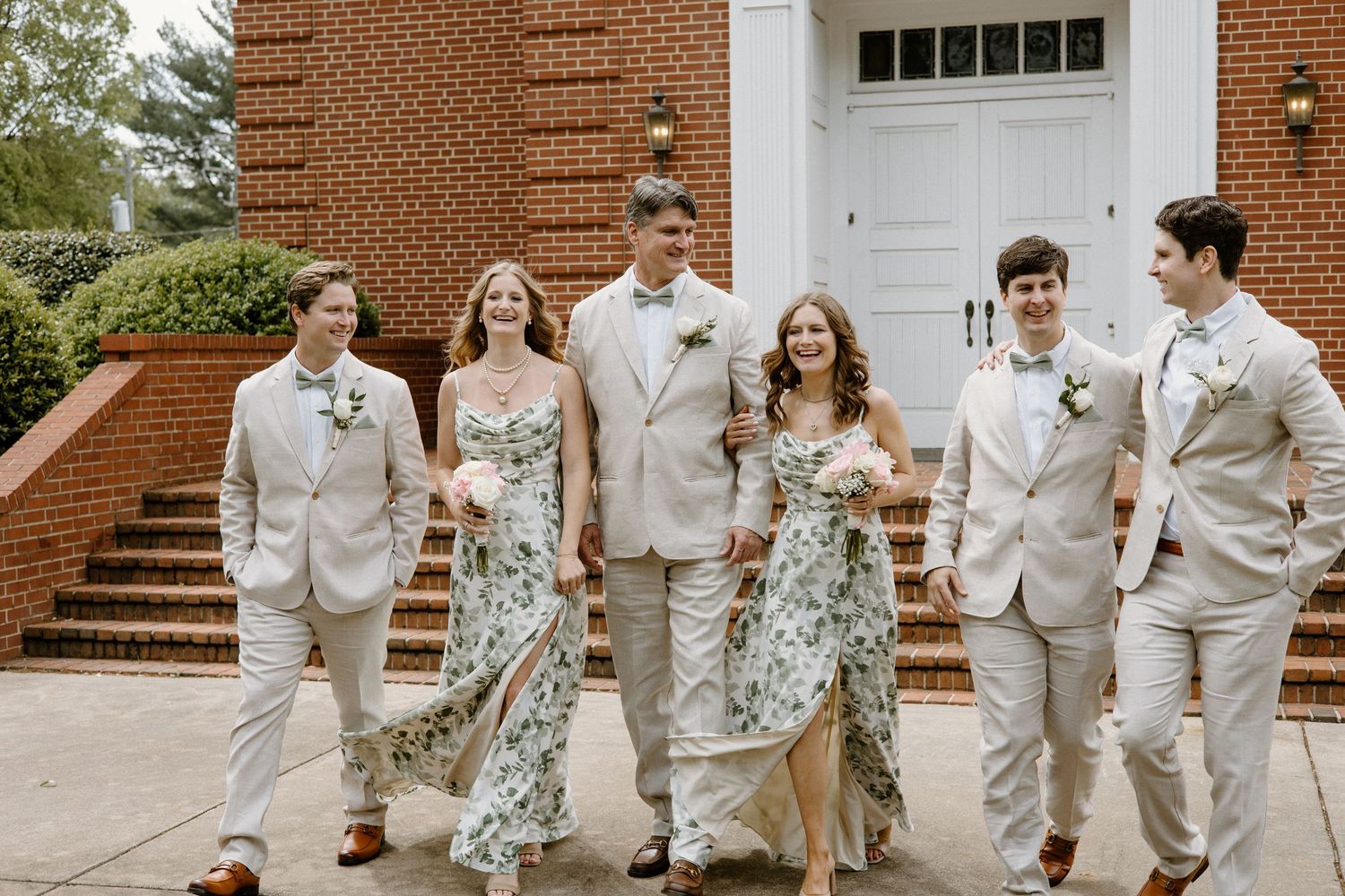 Wedding party in light colored suits and floral dresses poses together outside a brick church building.