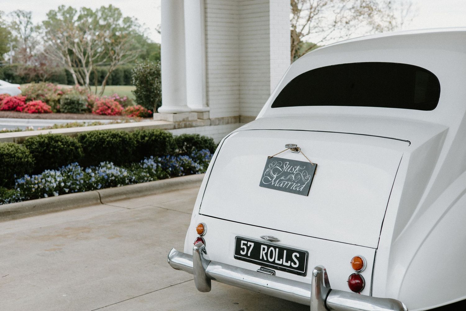 Rear view of a classic white vintage car parked in front of a columned building with manicured gardens.