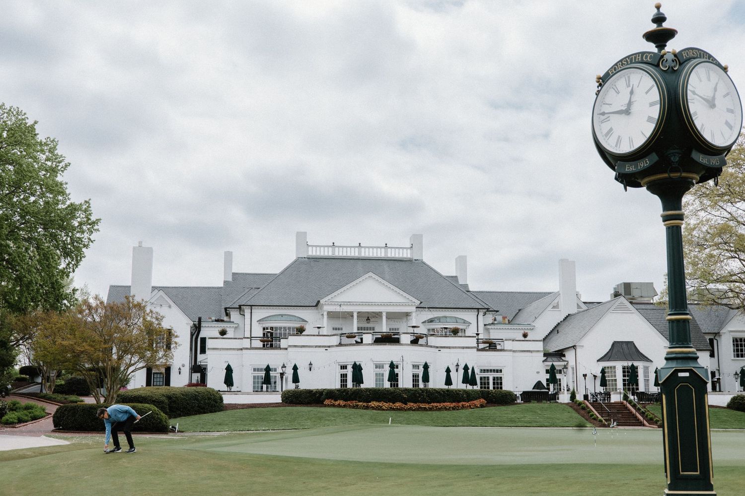 A white clubhouse with colonial architecture overlooks a manicured golf green with a vintage clock post in the foreground.