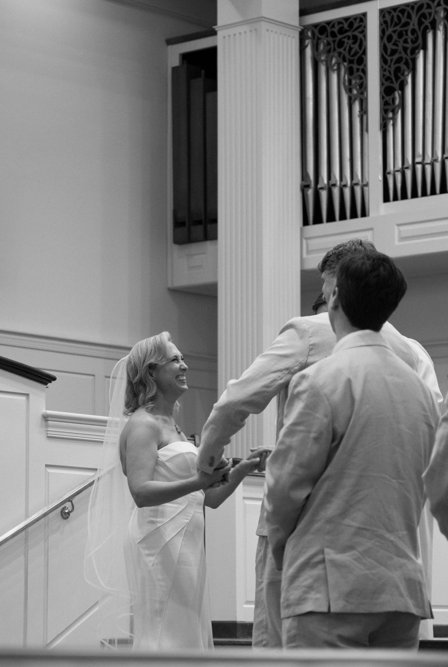 A bride and officiant stand together during a wedding ceremony with church organ pipes visible in the background.