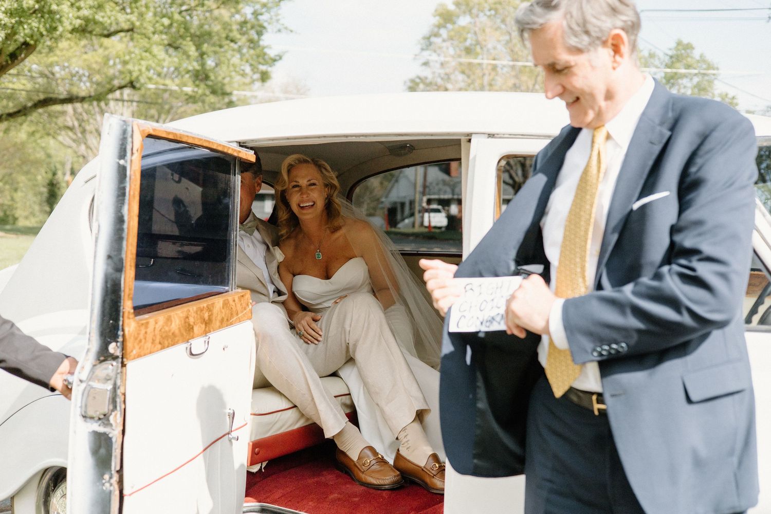 A bride in a white dress sits inside a vintage car as a man in a suit stands outside holding a gift.