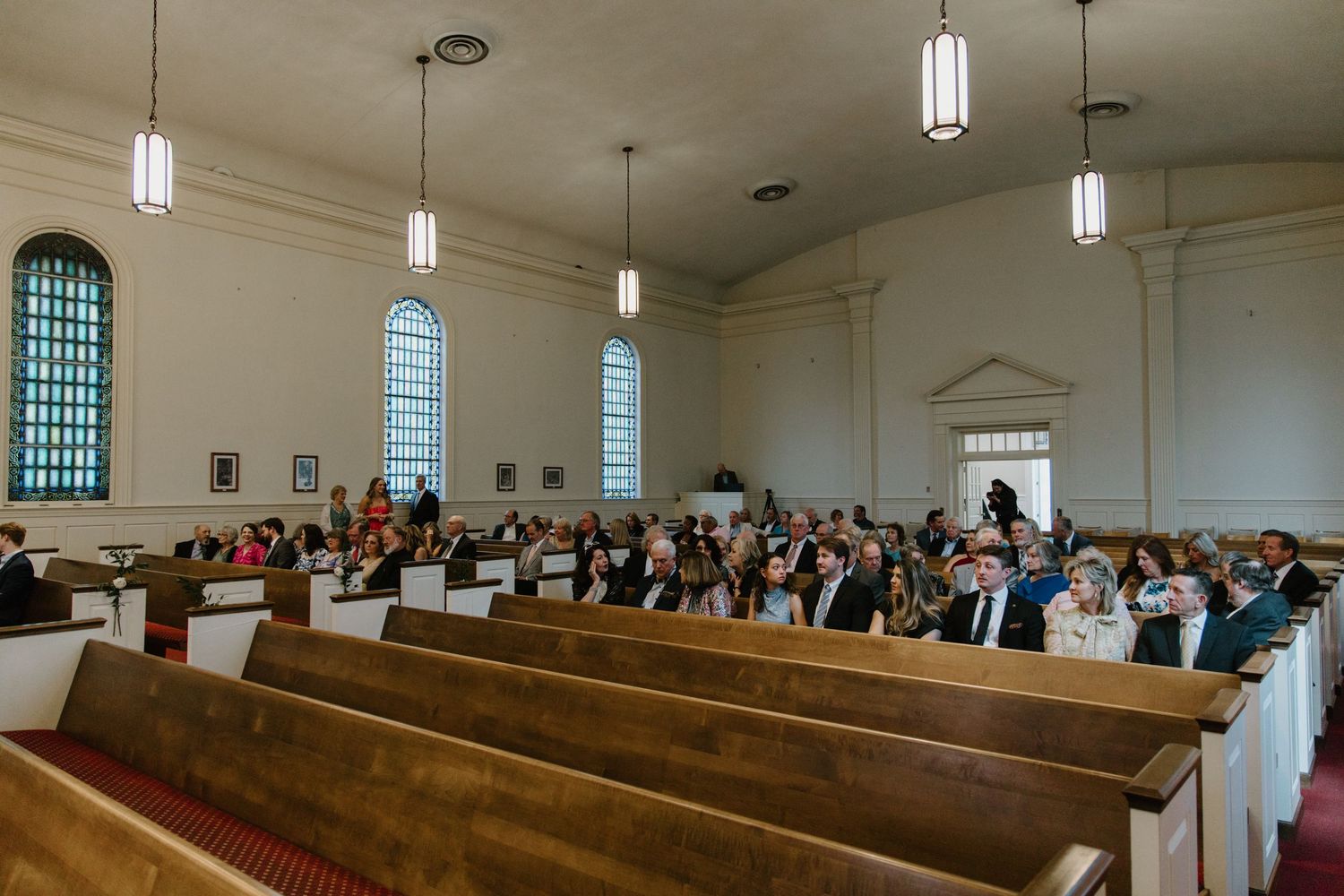 Wedding guests seated in wooden church pews with arched windows and hanging pendant lights illuminating the chapel interior.