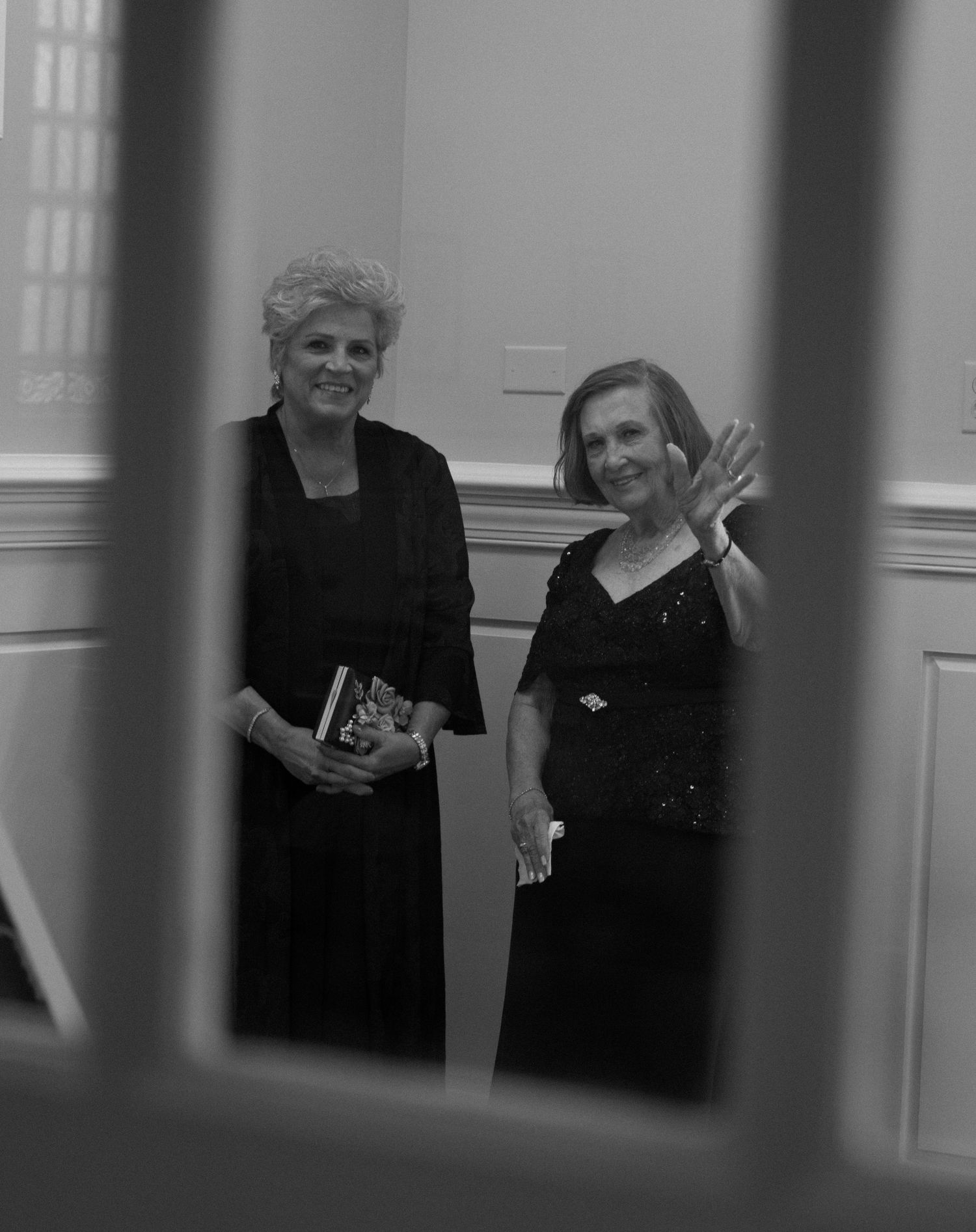 Two elegantly dressed women in black formal attire stand together in an ornate room.