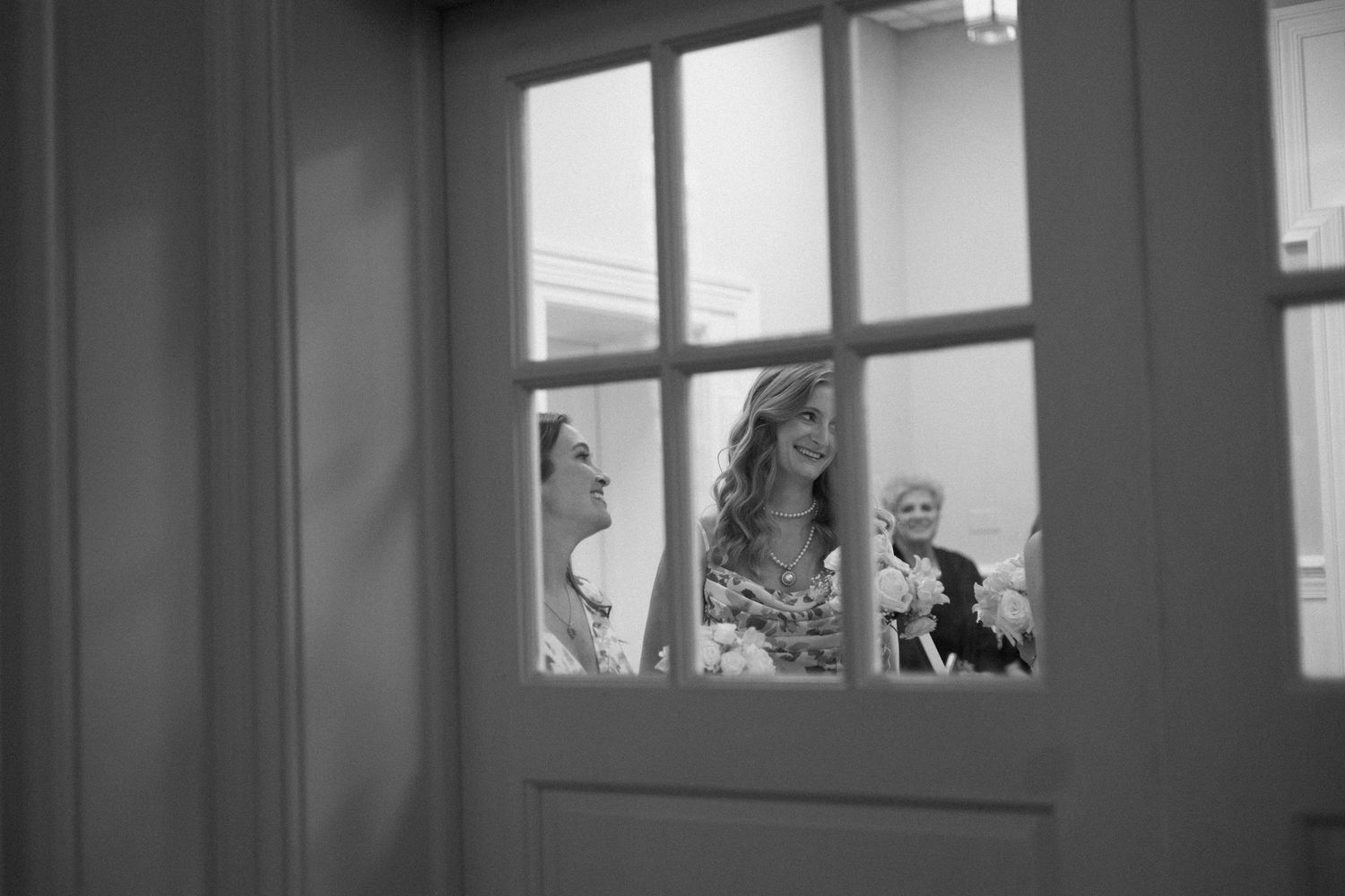 Black and white view through a window pane showing three people's silhouettes laughing and socializing indoors.
