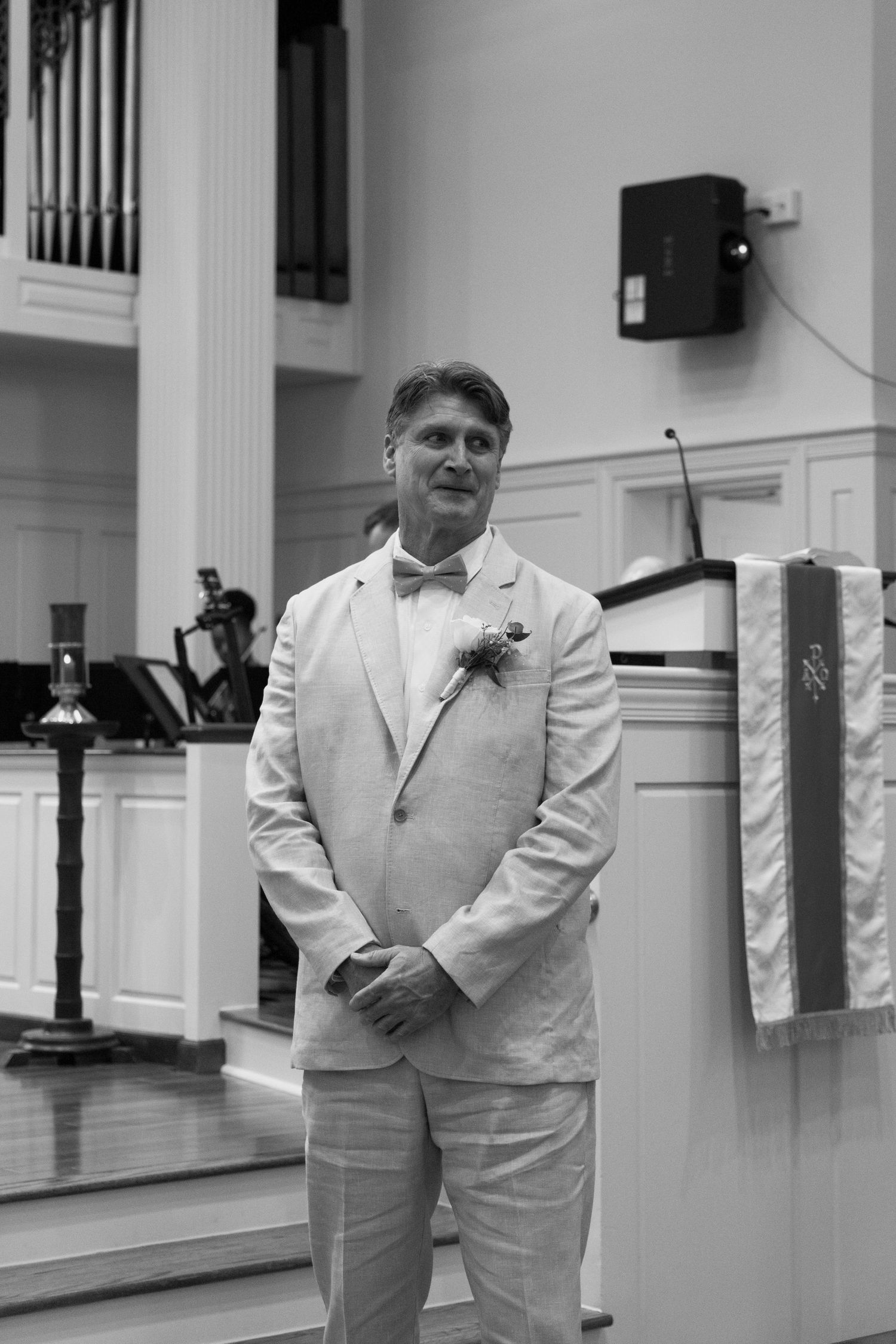A person in a white suit with a bow tie stands at the front of a church sanctuary in a black and white photograph.