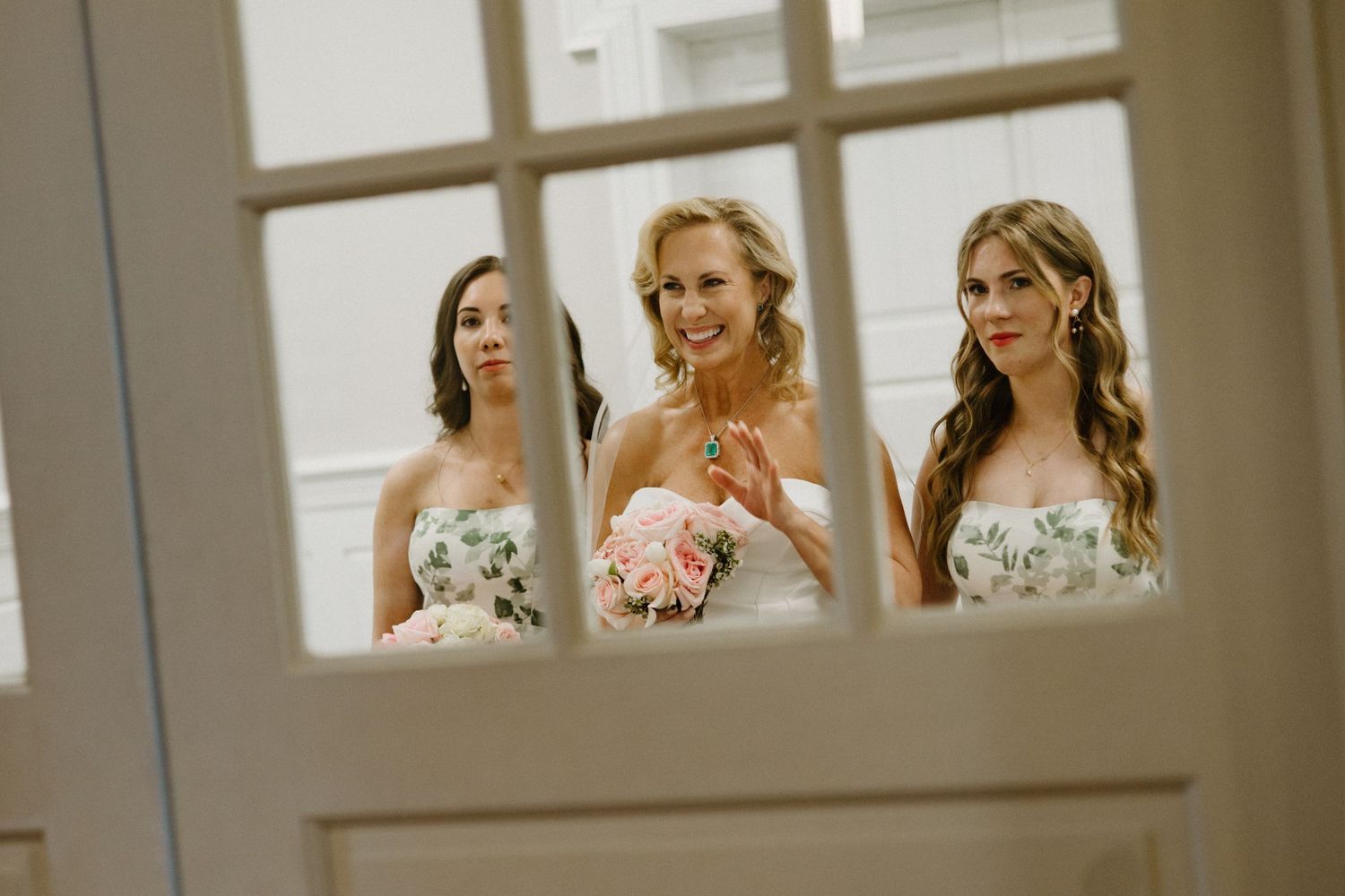 Three bridesmaids in strapless dresses with floral details peek through a window while holding bouquets.