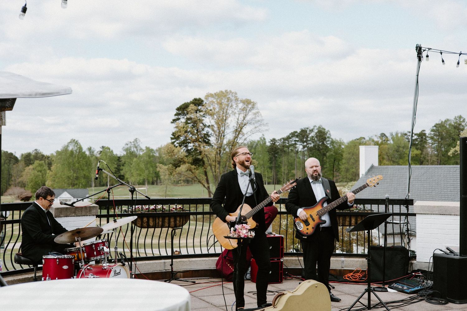 Musicians perform with guitars on an outdoor terrace during a daytime event with trees visible in the background.