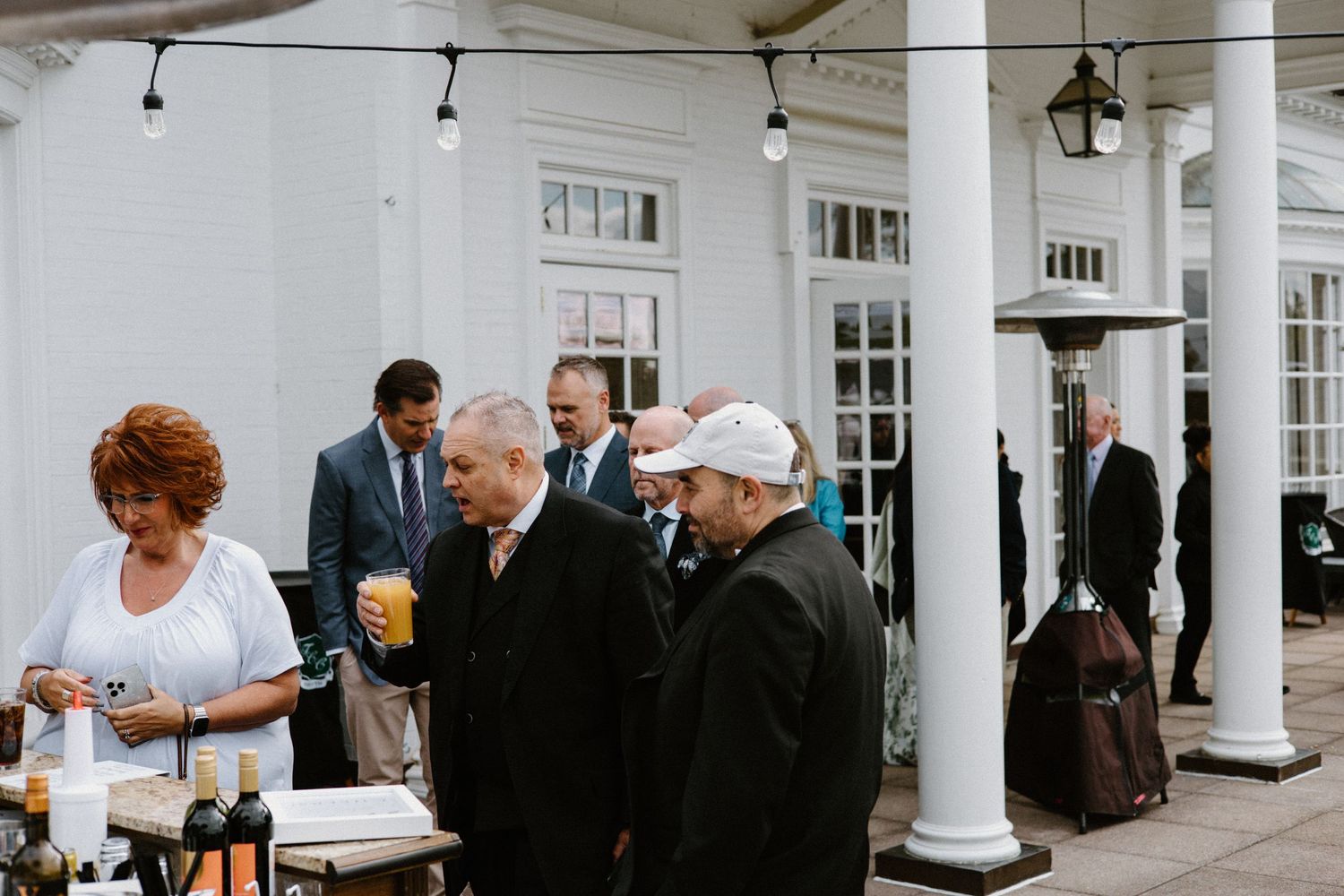 People gather at an elegant white columned porch during a social event with drinks being served.
